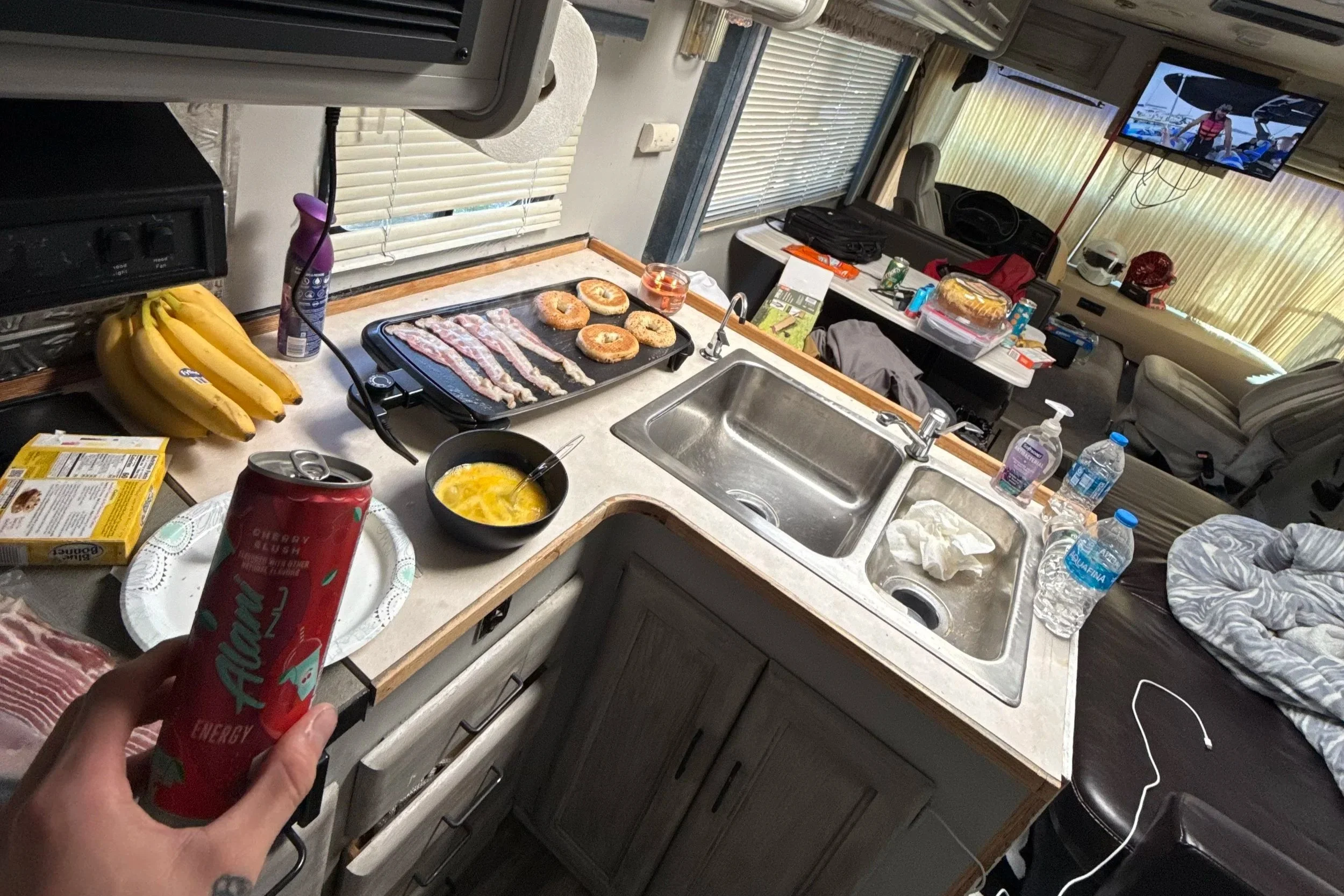 Kitchen countertop with bacon and donuts on a griddle, bananas, a bowl of scrambled eggs, and a hand holding a red energy drink can. Background shows a living area with a table, water bottles, and a TV.