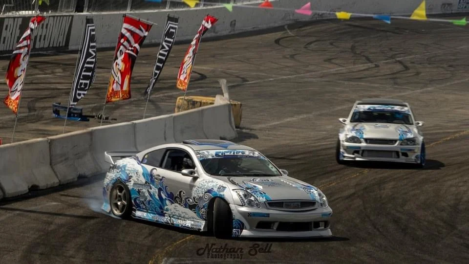 Two decorated race cars on a dirt track during a motorsport event, with colorful flags and banners in the background.