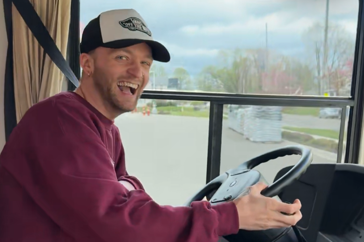 A man smiling and sitting at the steering wheel of a bus, wearing a black and white cap and a maroon sweatshirt.