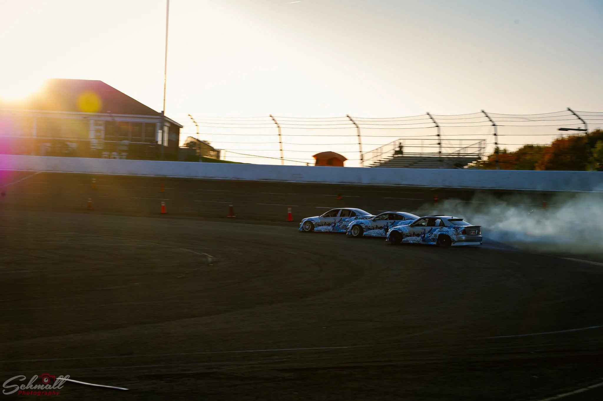 Three race cars drifting around a curve on a racetrack at sunset, with smoke trailing behind, orange cones on the track, and an empty grandstand in the background.