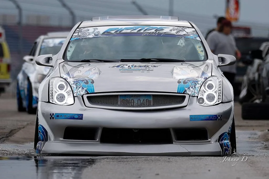 Silver racing car with blue and gray wave-inspired graphics parked at a racetrack, with other cars and people in the background.
