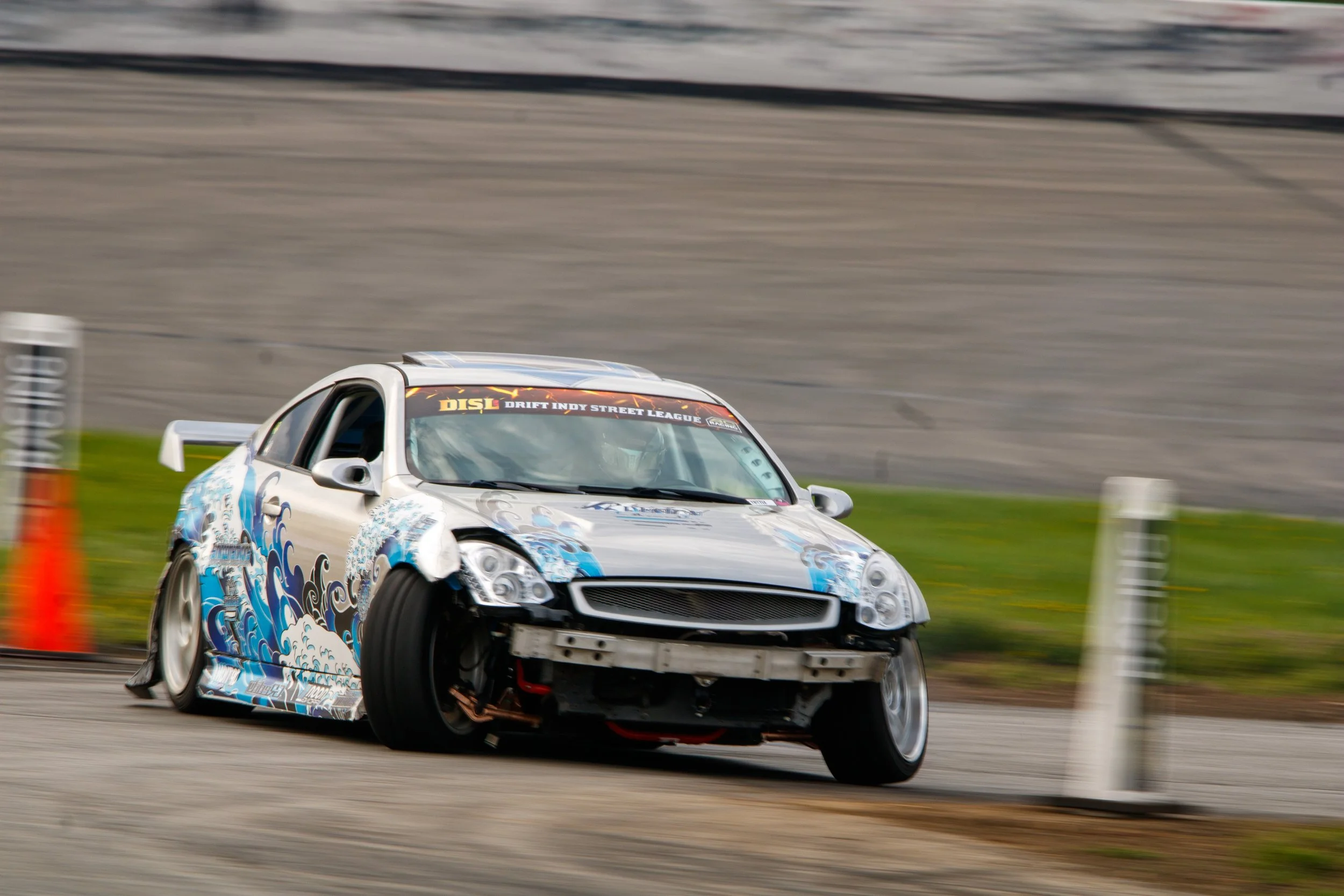 A silver race car with blue wave decals is drifting on a track, with the front-left wheel lifted off the ground. There are orange cones and blurred background indicating high speed.