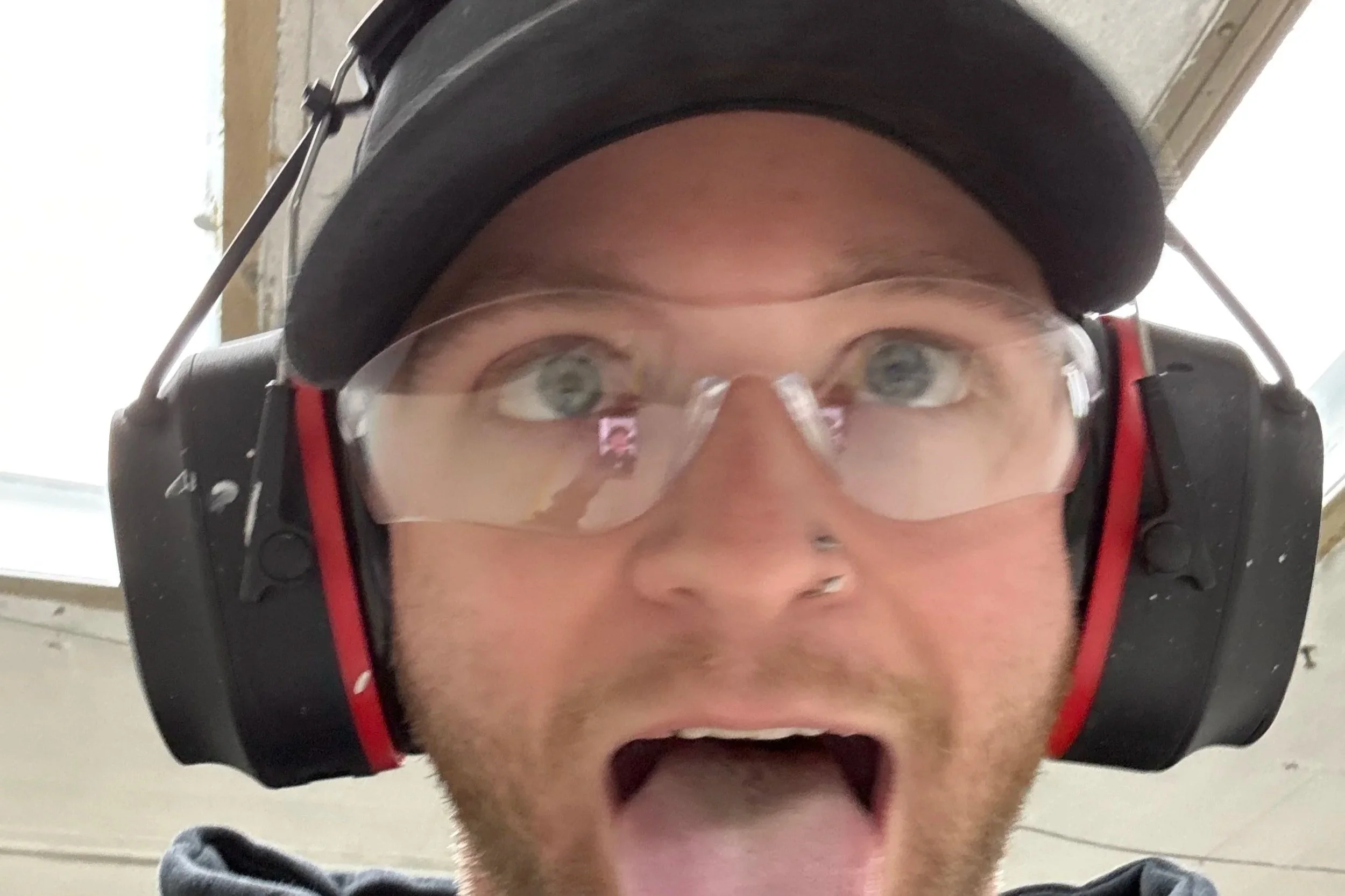 Close-up of a man wearing a black cap, safety glasses, and large noise-canceling headphones with a surprised or excited expression, mouth open, in an indoor setting.