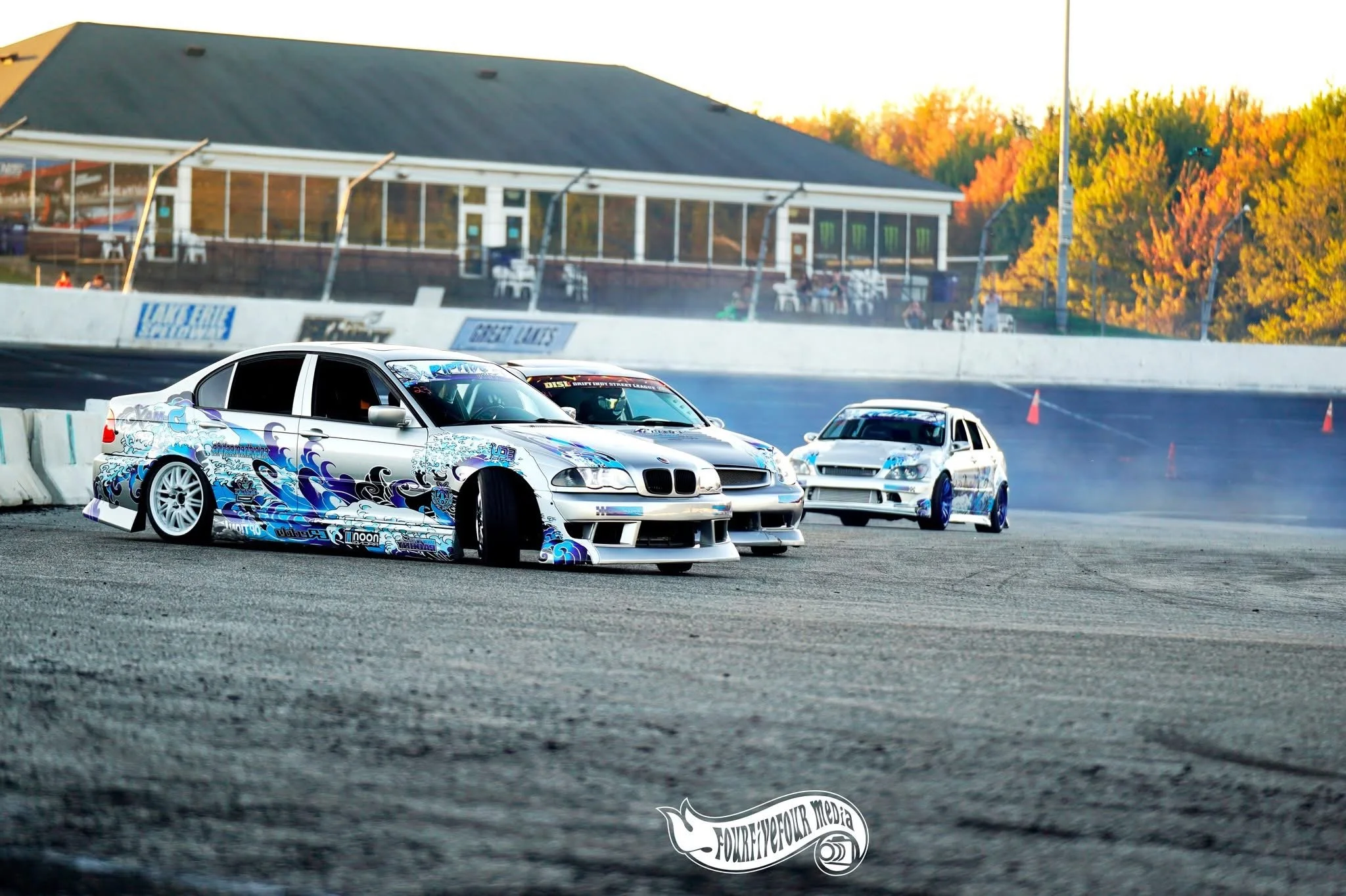 Three modified race cars on a dirt track, with the front car skidding and kicking up dust as they race during sunset.
