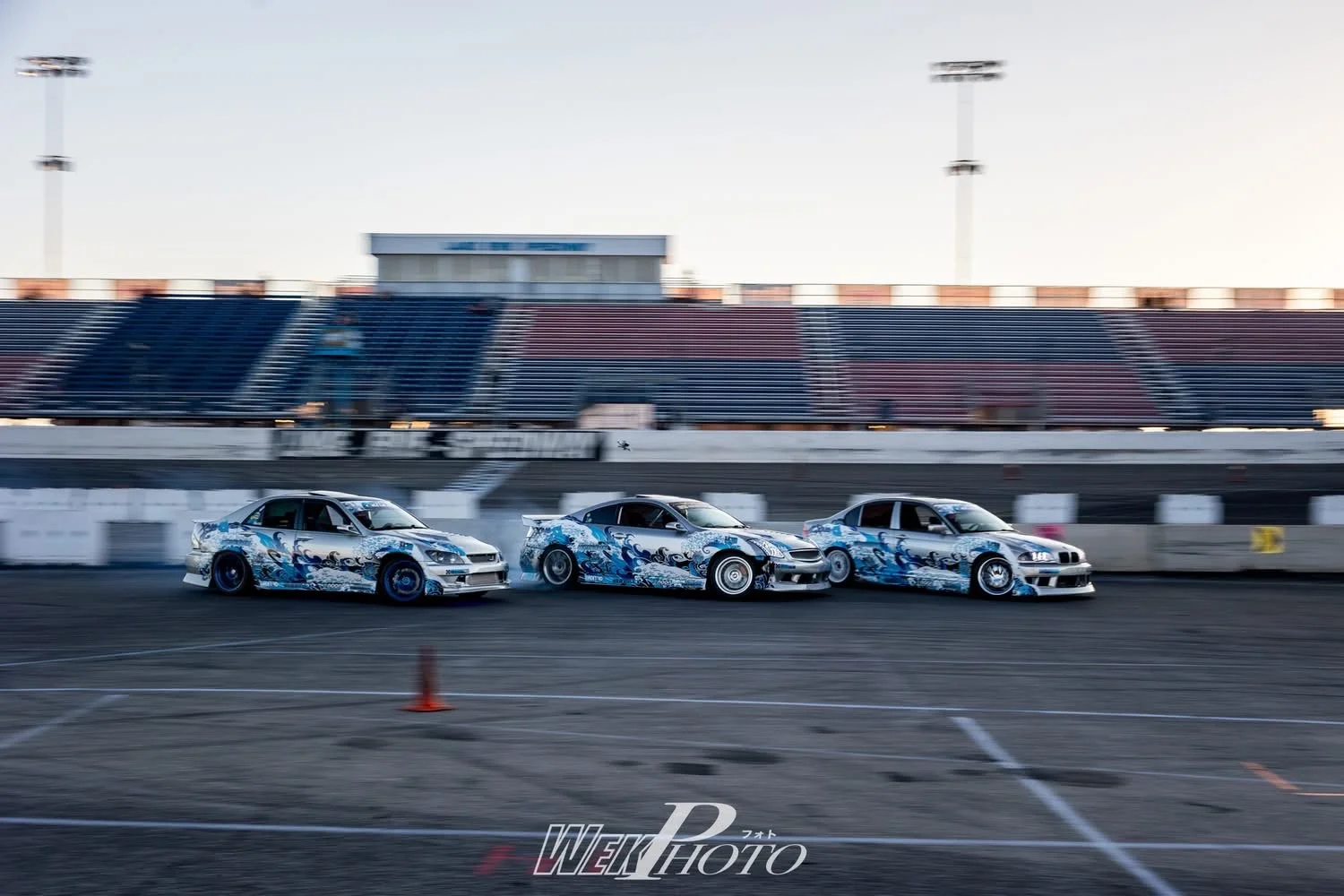 Three race cars with blue and white wave-themed graphics on a race track, blurred background of grandstands and bright sky at sunset.