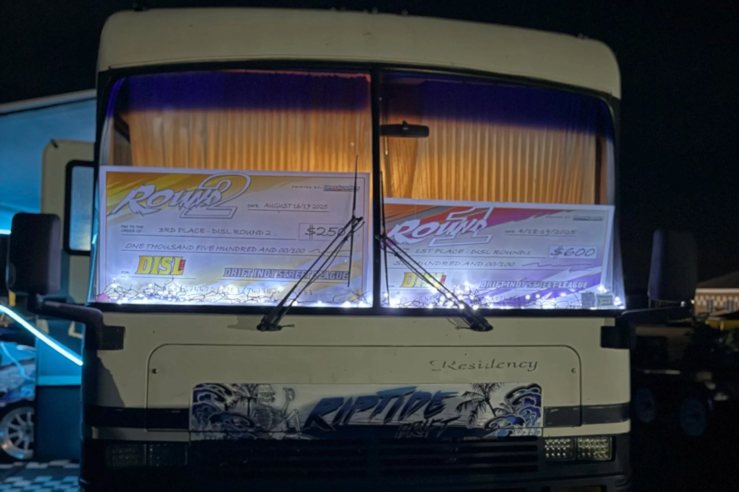 A bus at night with two large checks displayed in the front window, indicating race winnings for a drifting league, with the bus decorated with tropical-themed artwork and string lights.