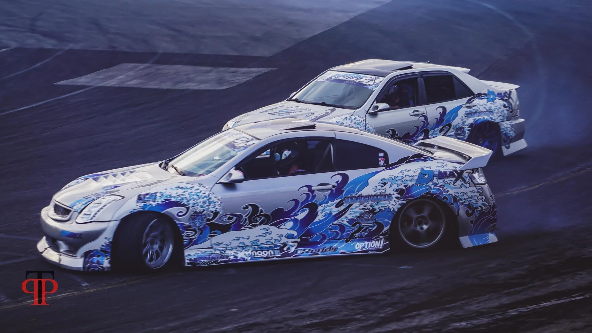 Two sports cars with blue and white wave-themed decals drifting on a race track.