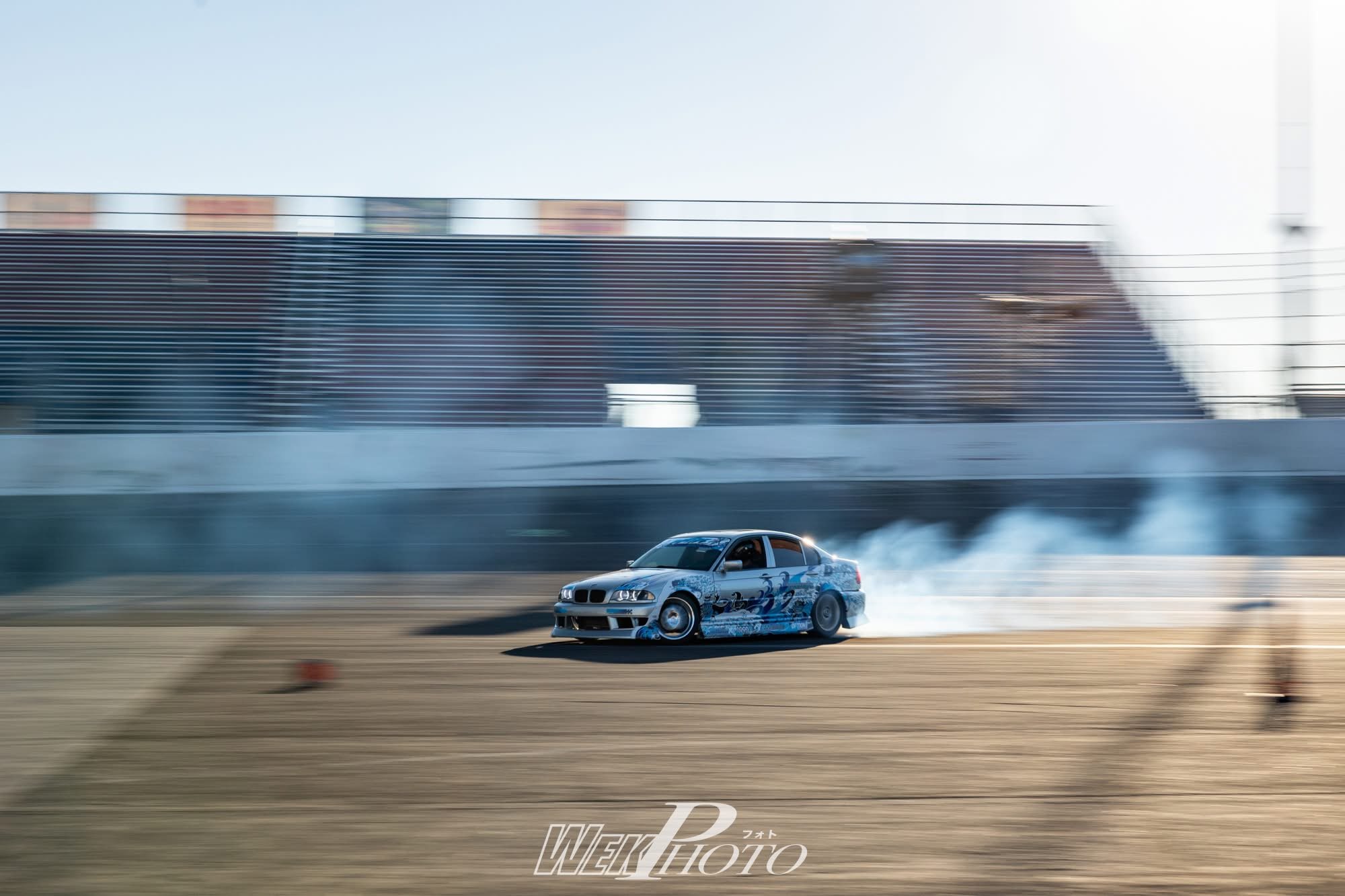 A racing car performs a drift on a race track, creating tire smoke with a blur background and a large building in the distance.