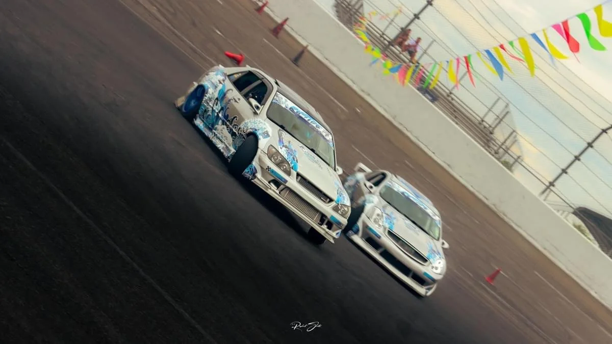 Two race cars with blue and gray livery on a dark race track, surrounded by colorful pennant banners and orange cones, with a spectator area above.
