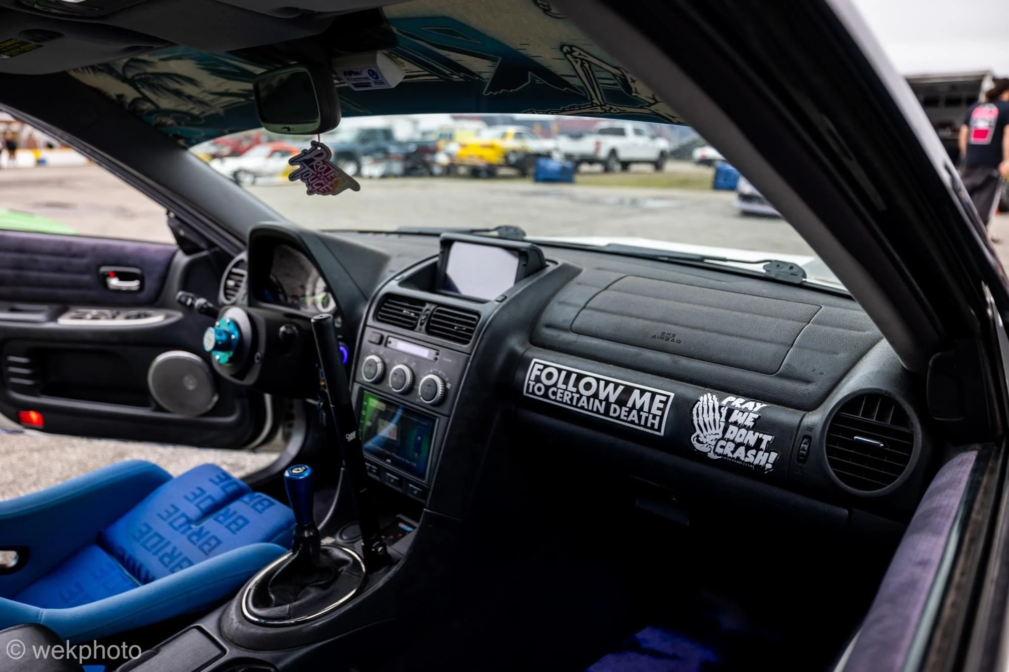 Interior of a car with racing modifications, including a blue racing seat, steering wheel, and gear shift, parked at a racing event or car meet with other cars and people in the background.