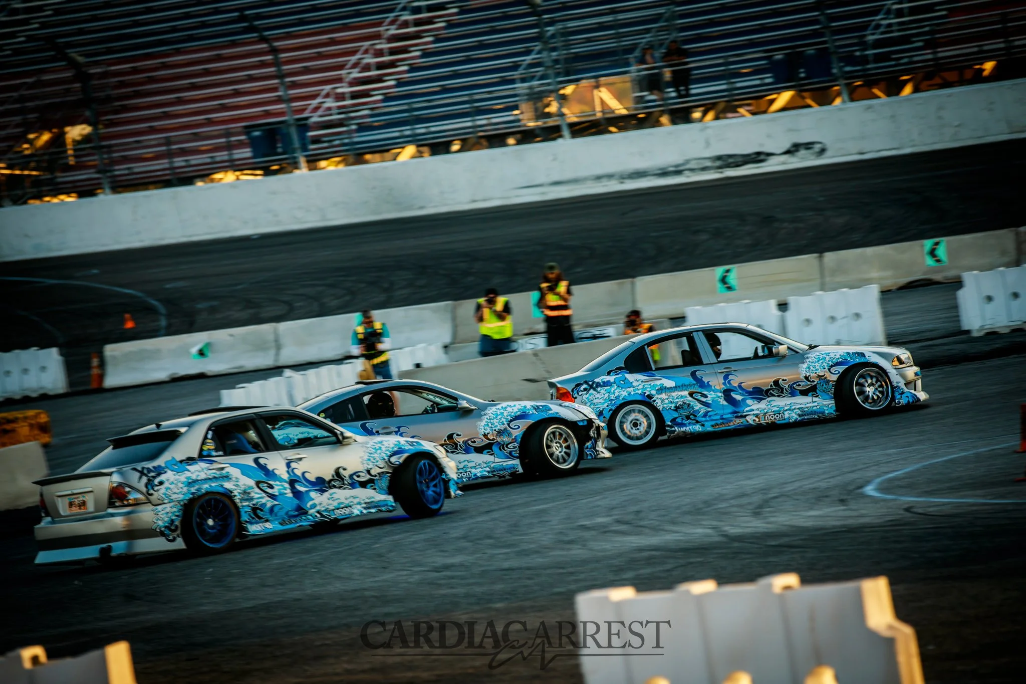 Two race cars with blue wave designs on the sides making a turn on a racetrack, with standing photographers and a multi-level grandstand in the background.