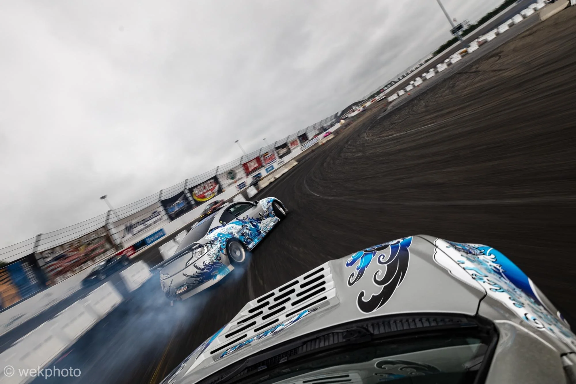 Two race cars on a dirt track, one in the foreground with a white, blue, and black design, and the other in the background with rust-colored paint, kicking up dust and smoke, under a cloudy sky.