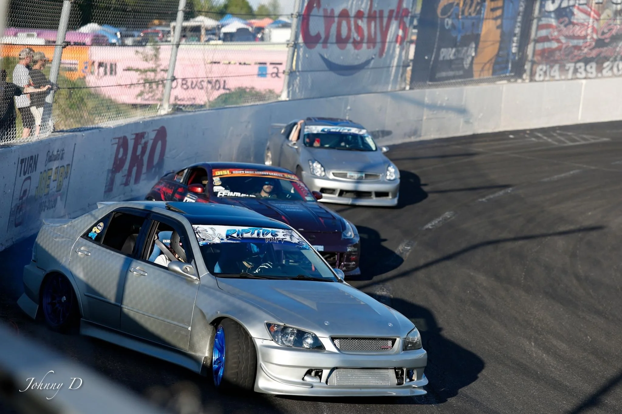 Four modified race cars on a racing track during a race, with spectators watching from the sidelines and colorful advertisements on the wall.