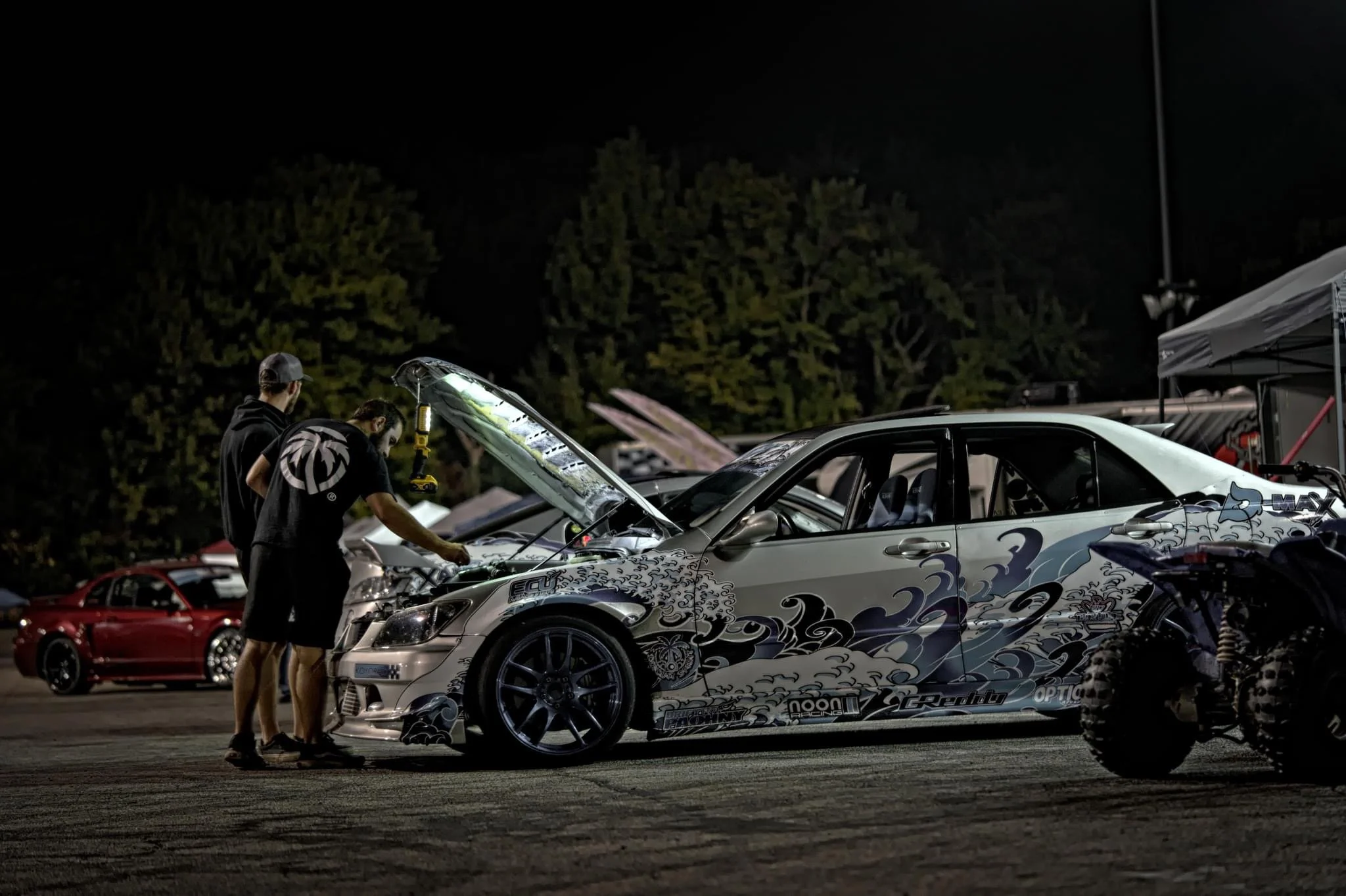 Two men working on a silver race car with a colorful wave design on the side at a night car event, with trees and another red car in the background.