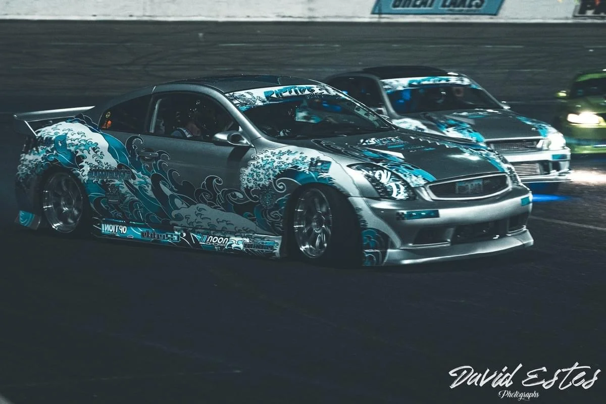 A silver race car with blue wave graphic and various decals, racing on a track at night with other cars visible in the background.