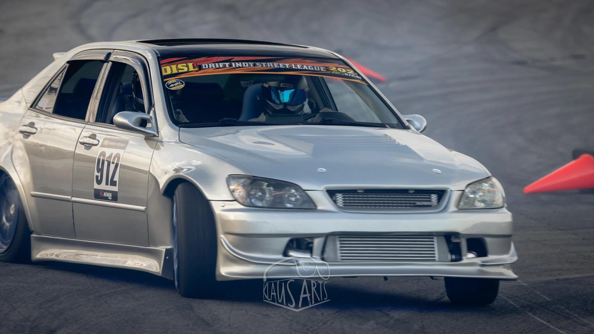 Silver race car with number 912 on the side, drifting on a dark track with a driver wearing a helmet.