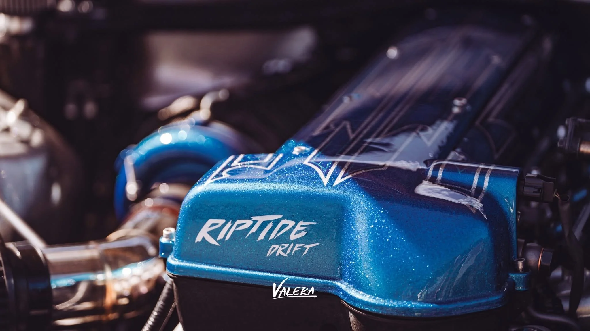 Close-up of a blue metallic car part with the words 'RIPTIDE DRIFT' written on it, with a background of mechanical components.