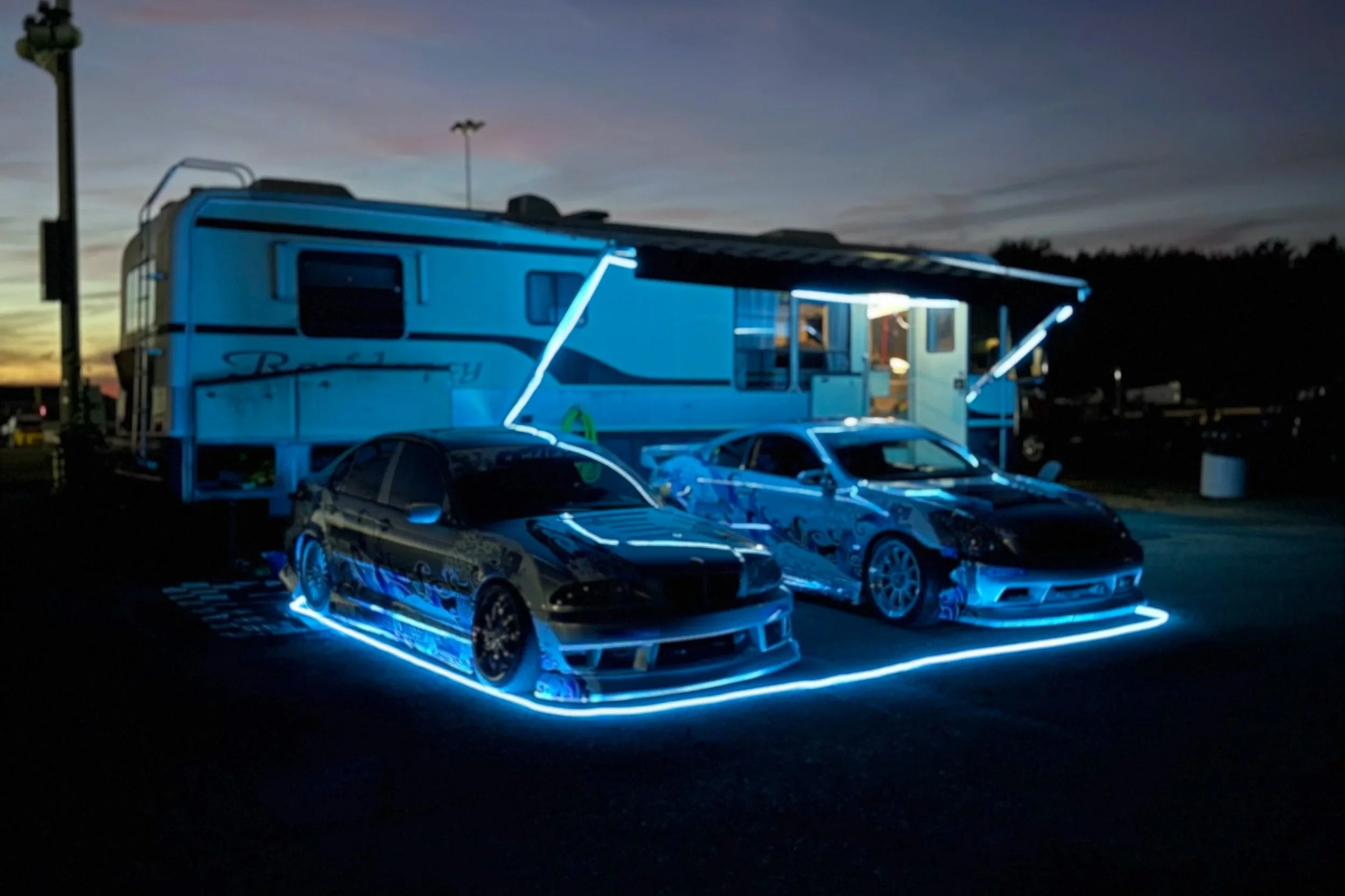 Two modified race cars with neon blue underglow lighting parked in front of a travel trailer during dusk at a race track.