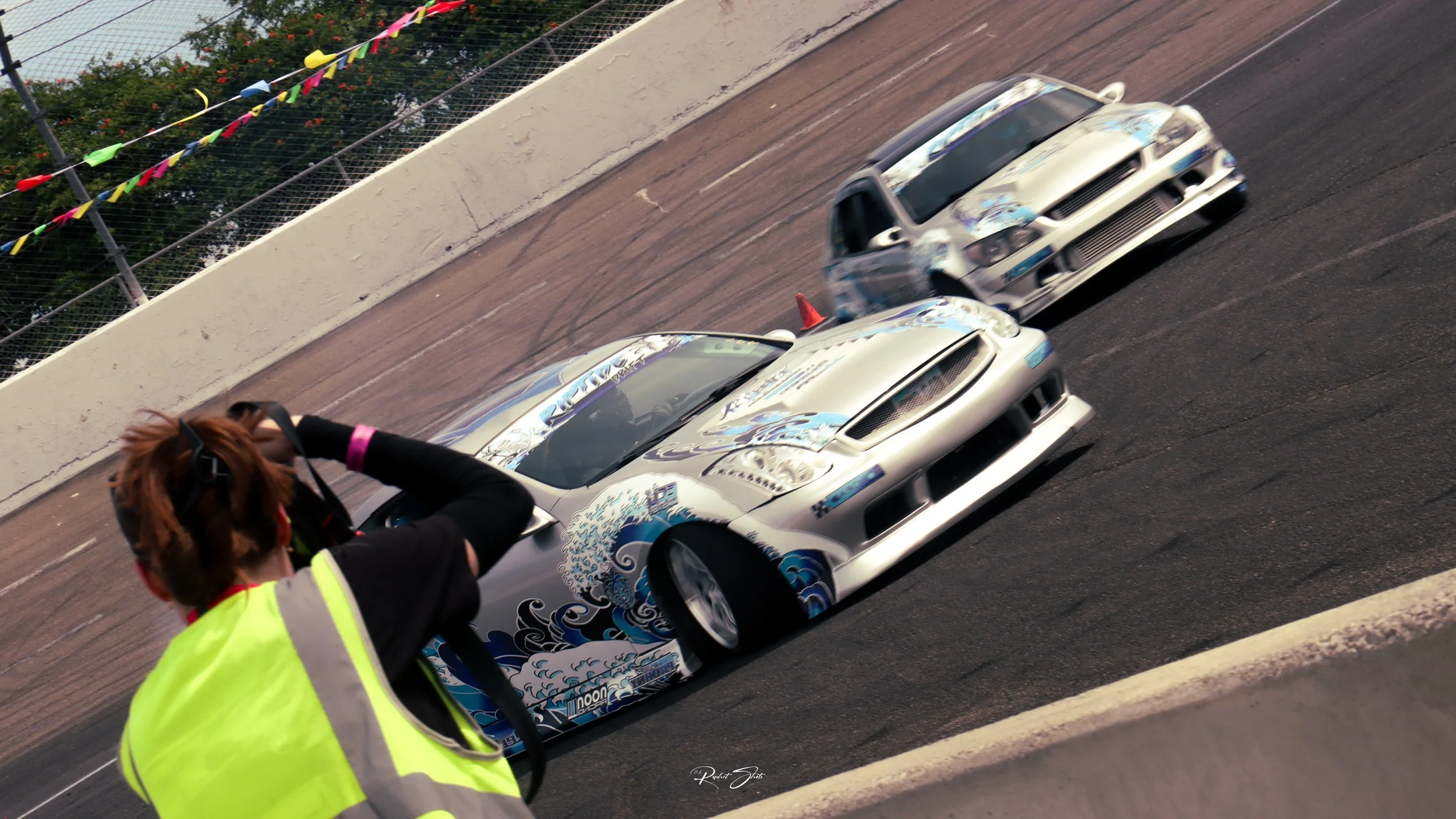A photographer wearing a neon yellow safety vest takes a picture of two race cars on a track. The cars are silver with blue wave designs, moving at high speed.
