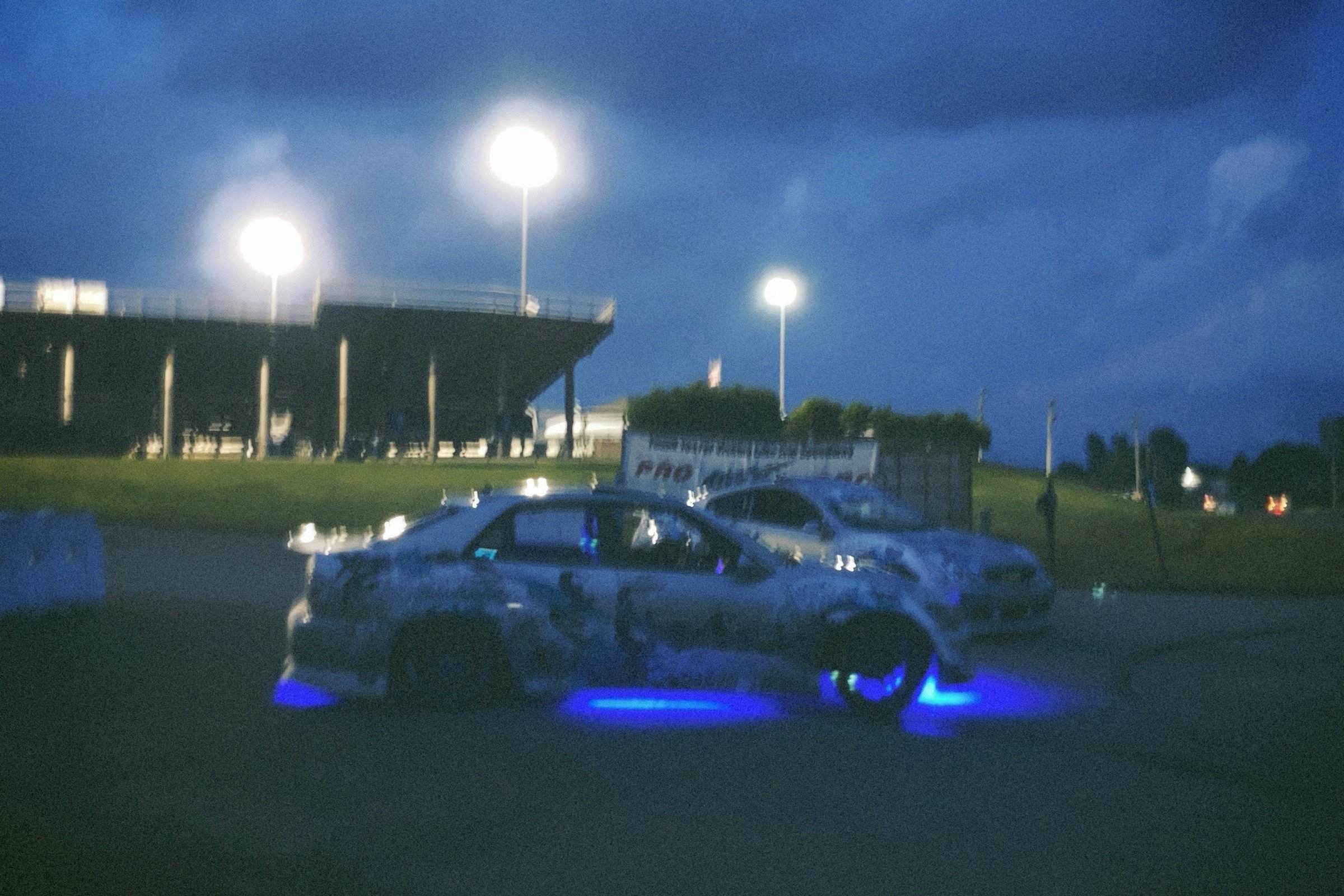 Nighttime scene with illuminated race cars on a track, surrounded by grass and bright stadium lights.