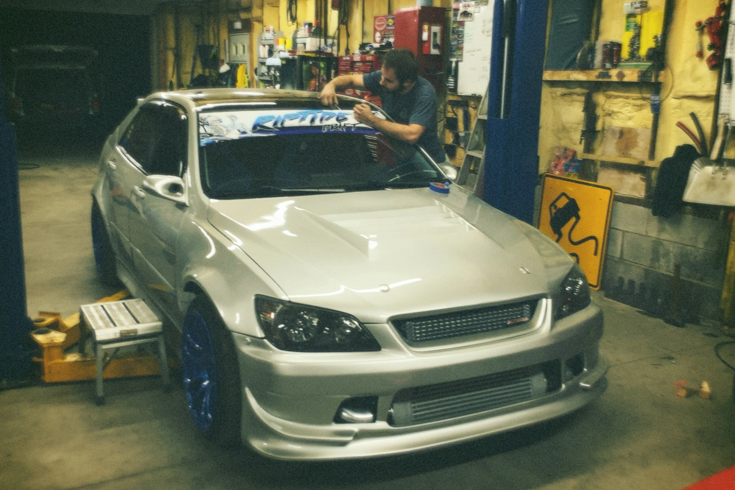 A man working on a silver race car inside a cluttered garage. The car has decals on the windshield, blue wheels, and a large front grille. Shelves with tools and equipment are visible in the background.