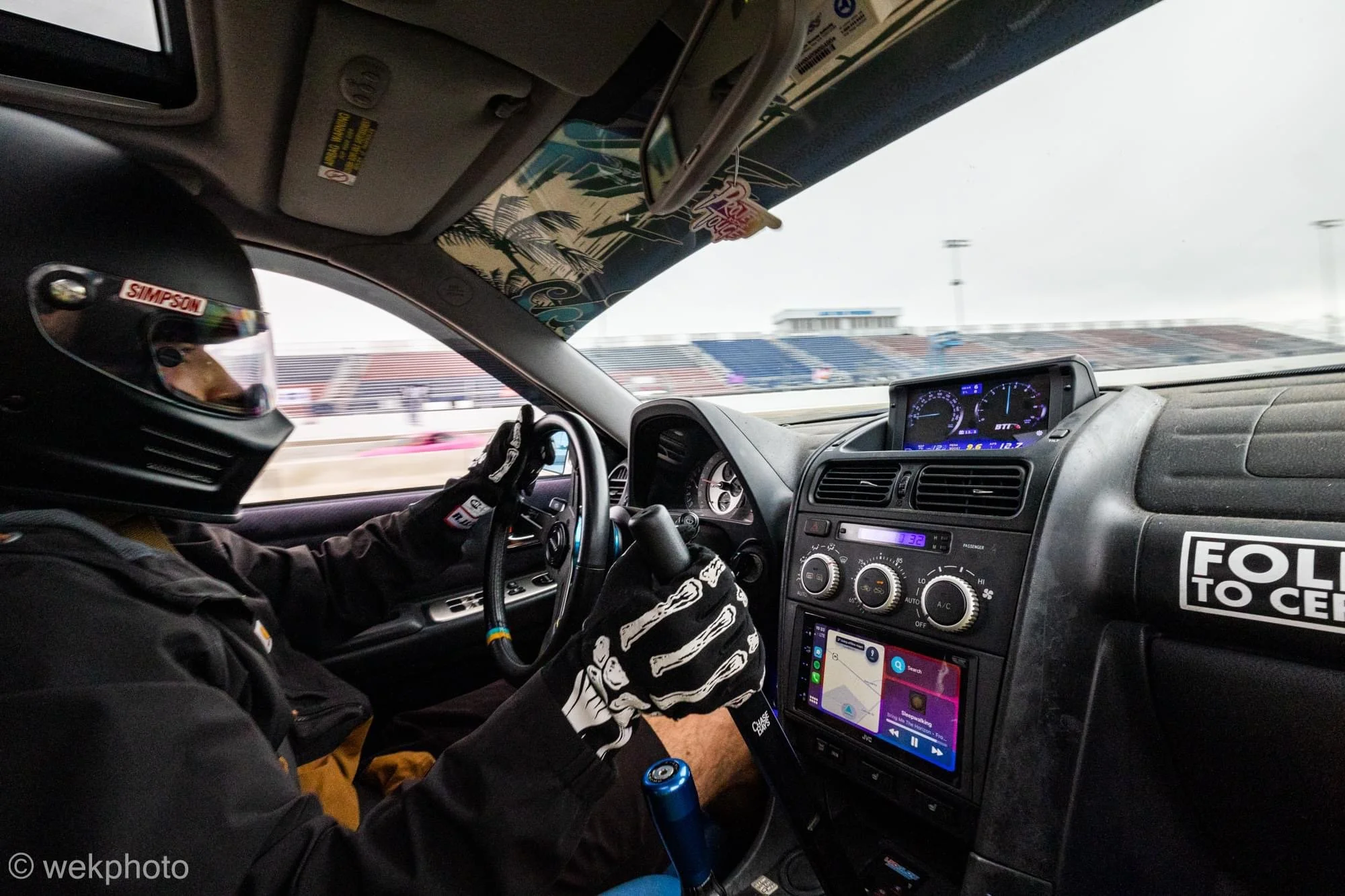 Inside a race car with a driver wearing a helmet and racing gloves, on a racetrack with stands in the background.