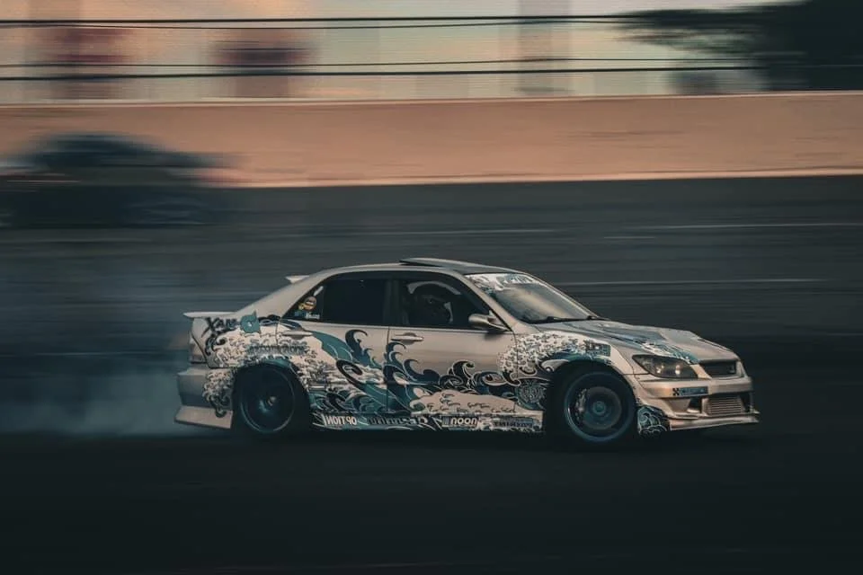 A silver race car with blue and black wave-themed graphics on its body speeding on a race track, creating tire smoke.