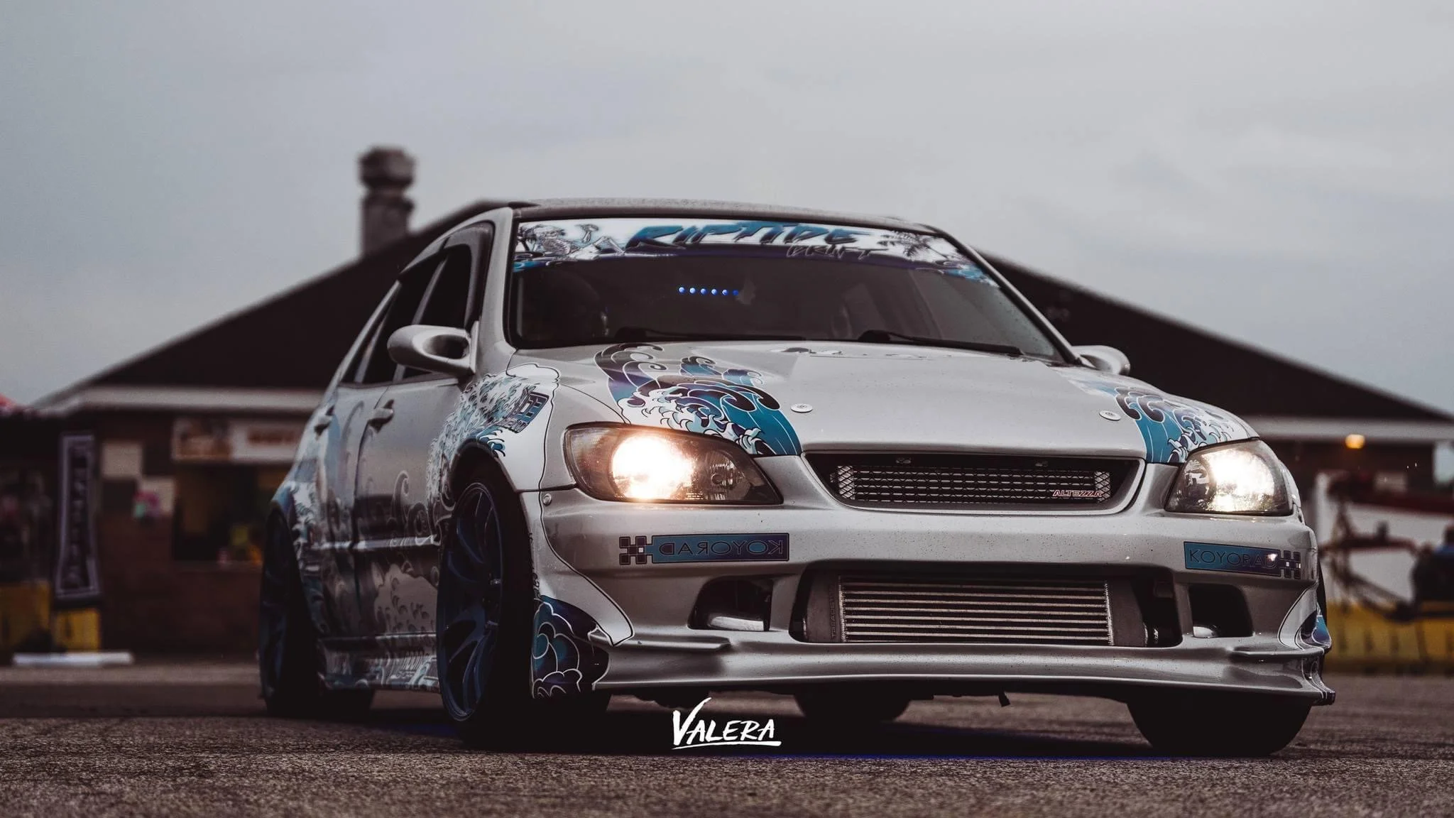 A customized silver race car with blue and black graphic decals park on a race track, with a building and cloudy sky in the background.