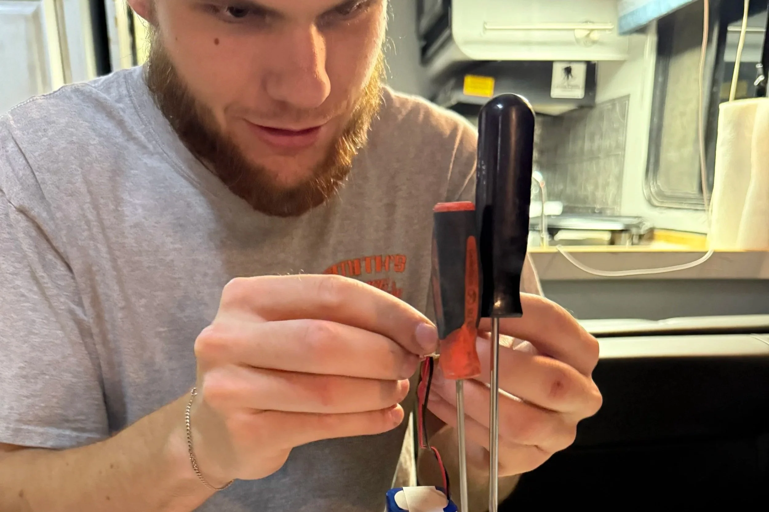 Man with a beard working on electronic components with a multimeter inside a kitchen or workshop.