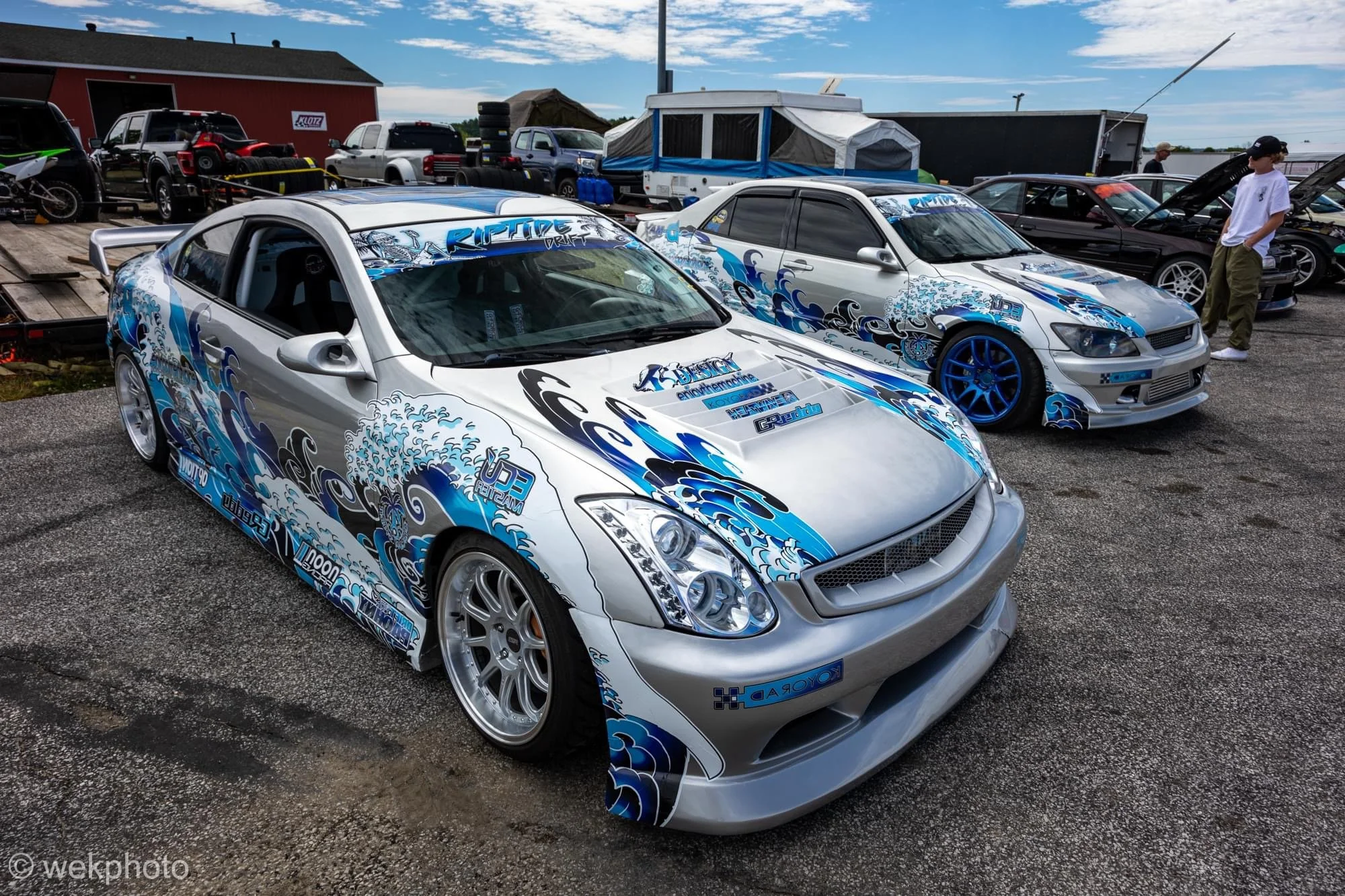 Two modified sports cars with blue and white wave-themed decals parked side by side at a car event, with other vehicles and people in the background.