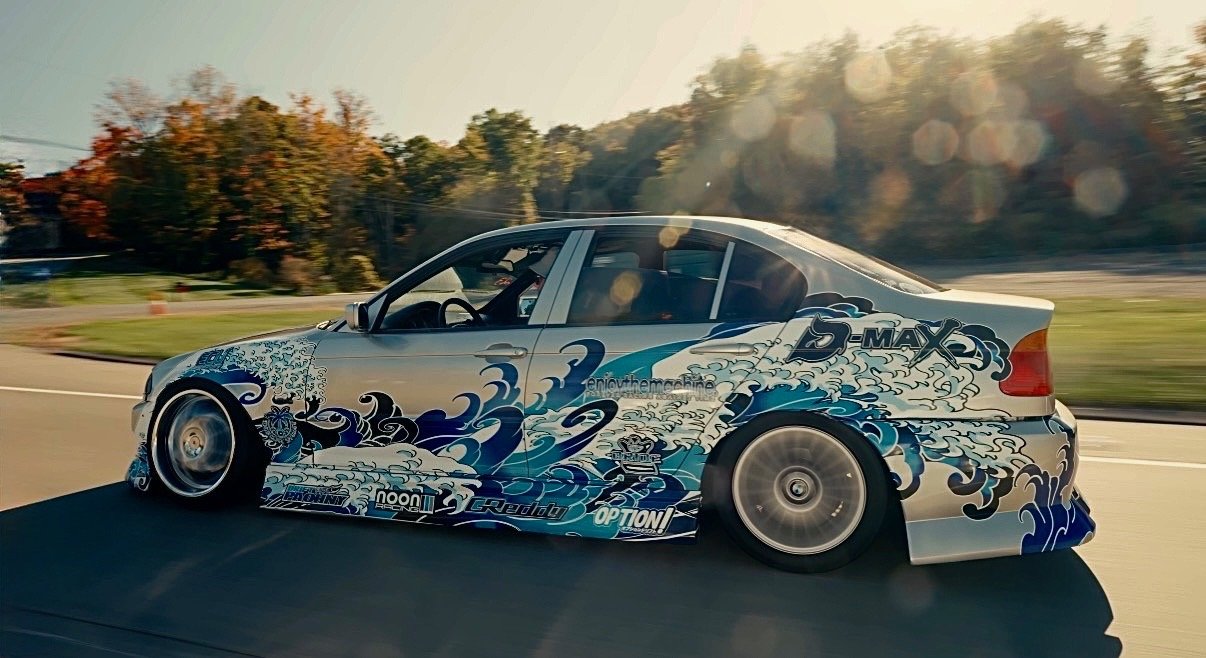 A silver racing car with blue and black wave designs and sponsor decals, driving on a highway with trees and a clear sky in the background.