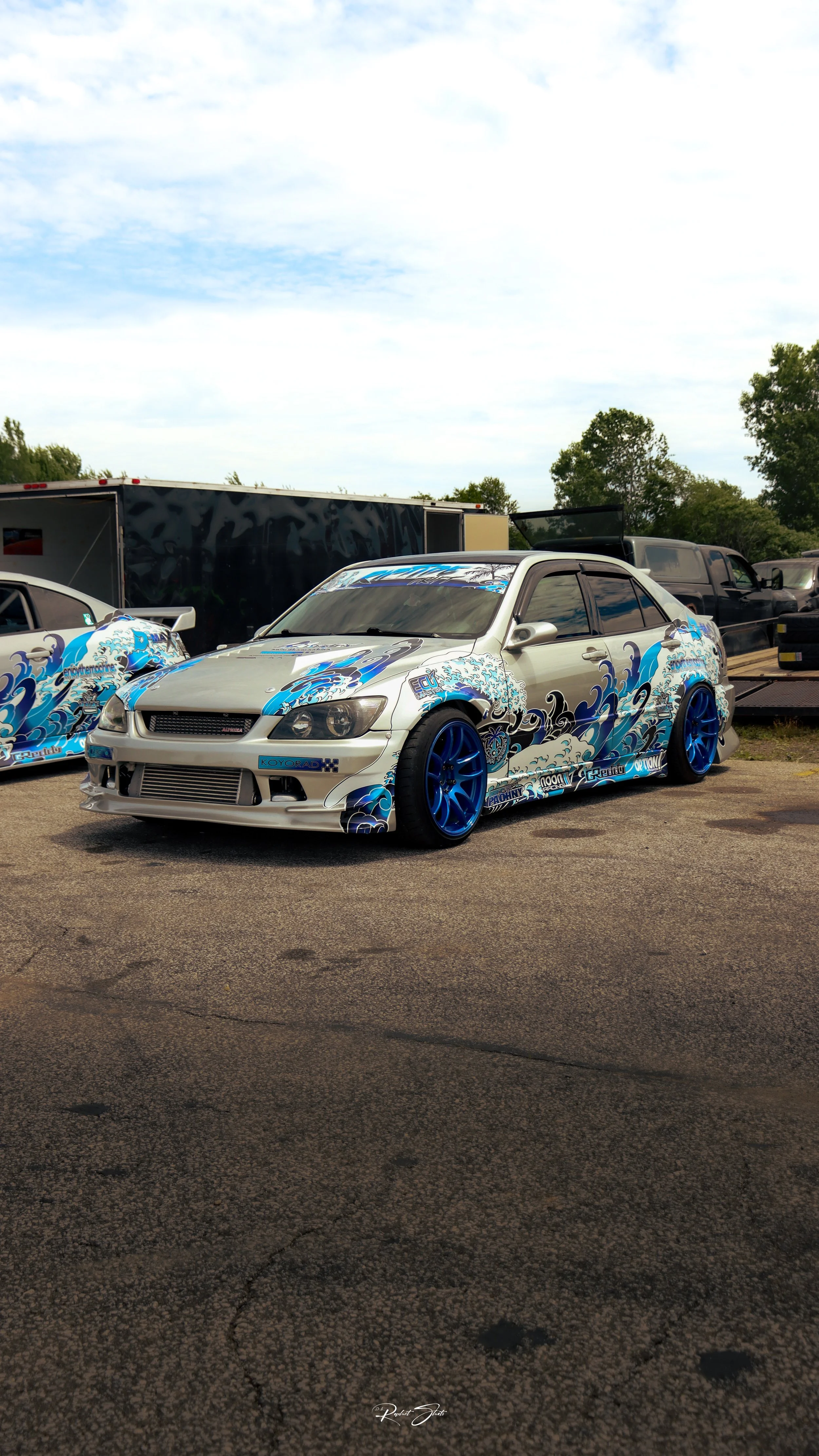 A modified white car with blue and black graphic wrap featuring wave designs, blue rims, parked on asphalt.