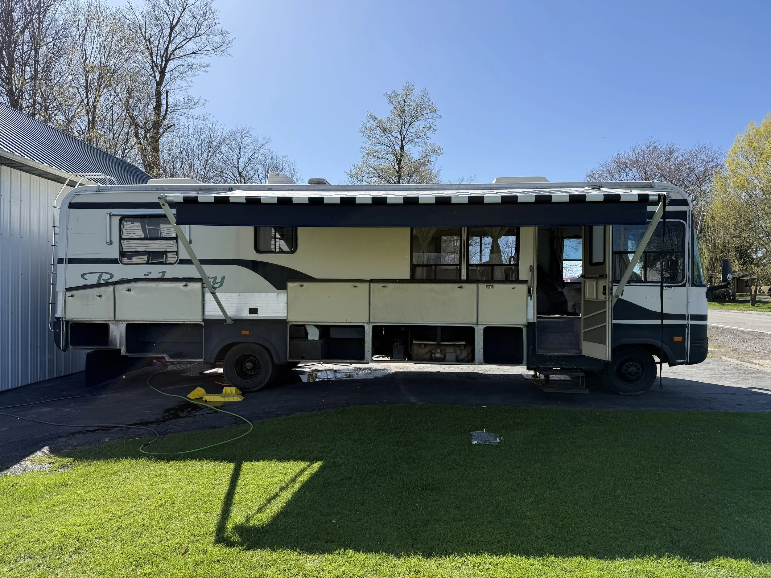Old, white and black RV parked on a paved driveway with a grass lawn in front, partially open door, window curtains, and an extended awning, under a blue sky, with leafless trees and a metal building in the background.