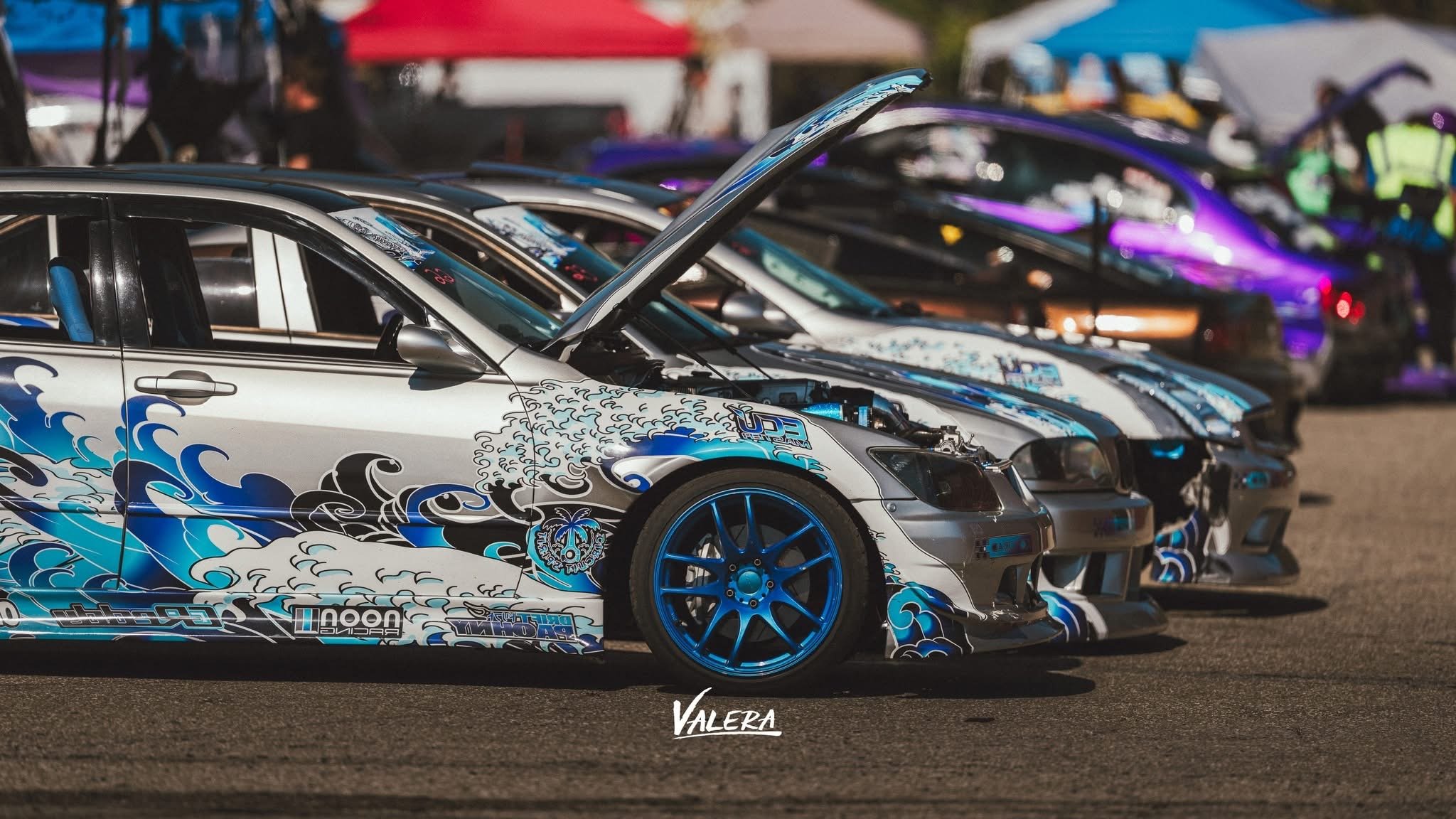 A row of modified race cars with colorful liveries parked at a racing event. The image prominently features a silver car with blue and black wave graphics and blue rims with a lowered stance. The cars are lined up side by side, some with open hoods, and are set against a background of other vehicles and event attendees.