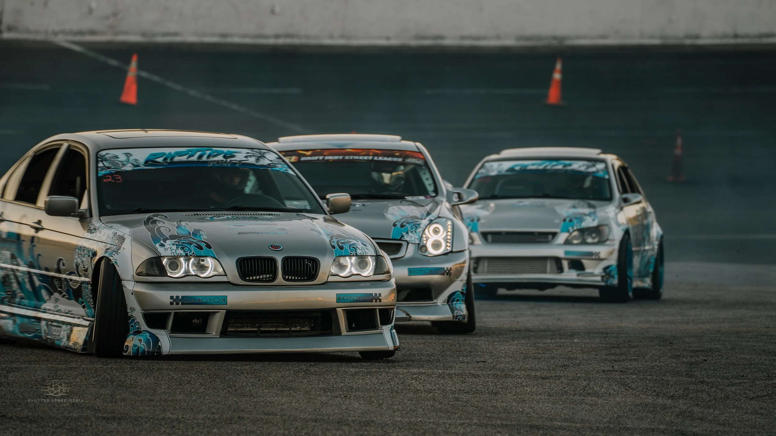 Three modified race cars in a drifting competition on a racetrack, with orange cones in the background.