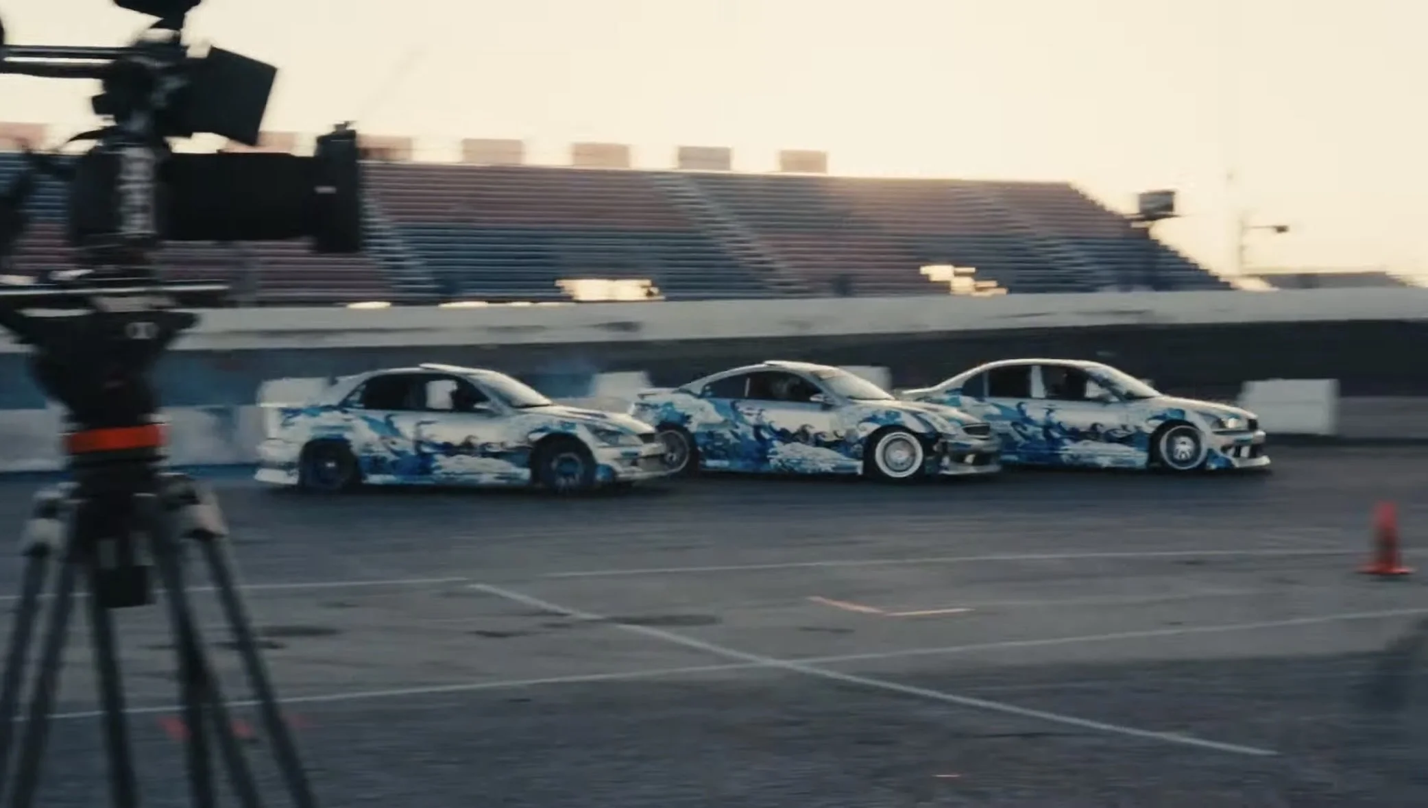 Three race cars with blue and white designs on a racetrack, with a camera on a tripod in the foreground and empty bleachers in the background.