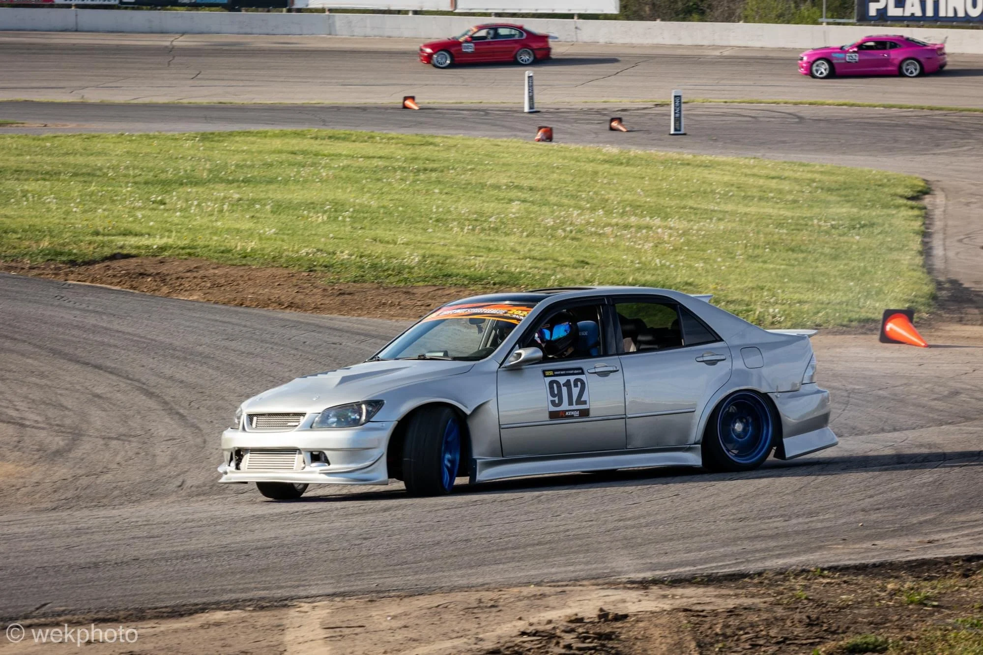 A silver race car with the number 912 is drifting around a track corner, with a driver wearing a helmet visible through the window. There are orange cones and a grassy area next to the track, and two other race cars are visible in the background on t