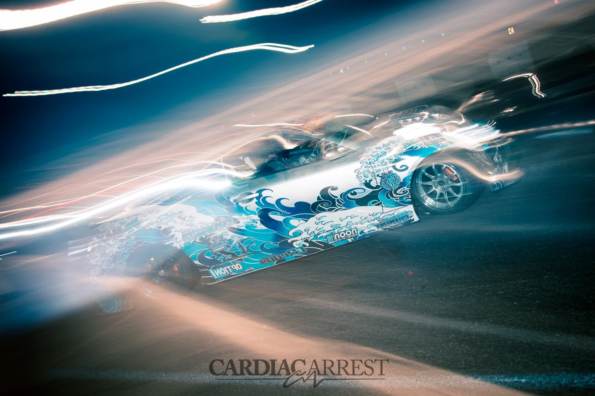 A race car with a blue and white wave design speeding on a track at night, with motion blur and light streaks around it.