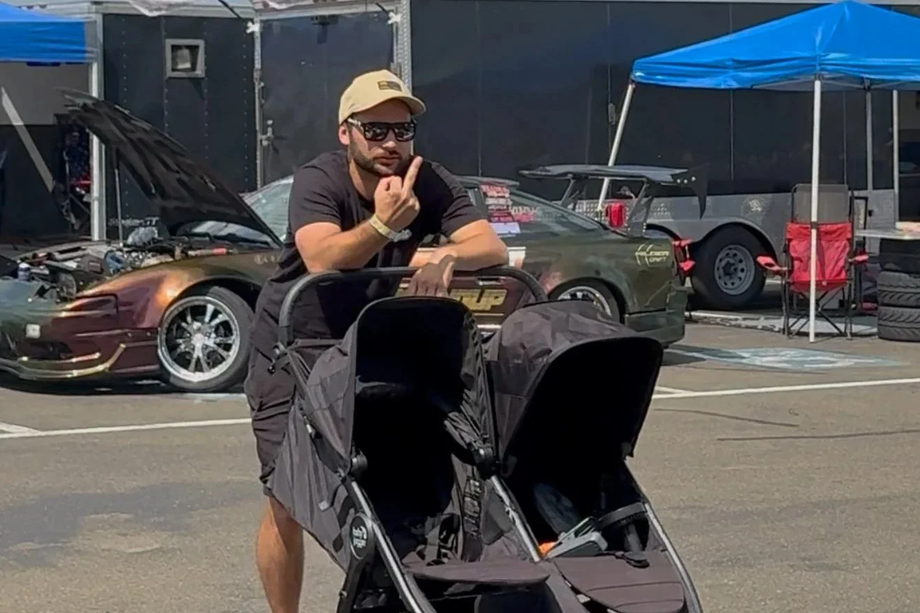 Man with beard and sunglasses, wearing a tan cap and black shirt, posing with peace sign near a stroller, behind a racing car in a paddock area at a motorsport event.