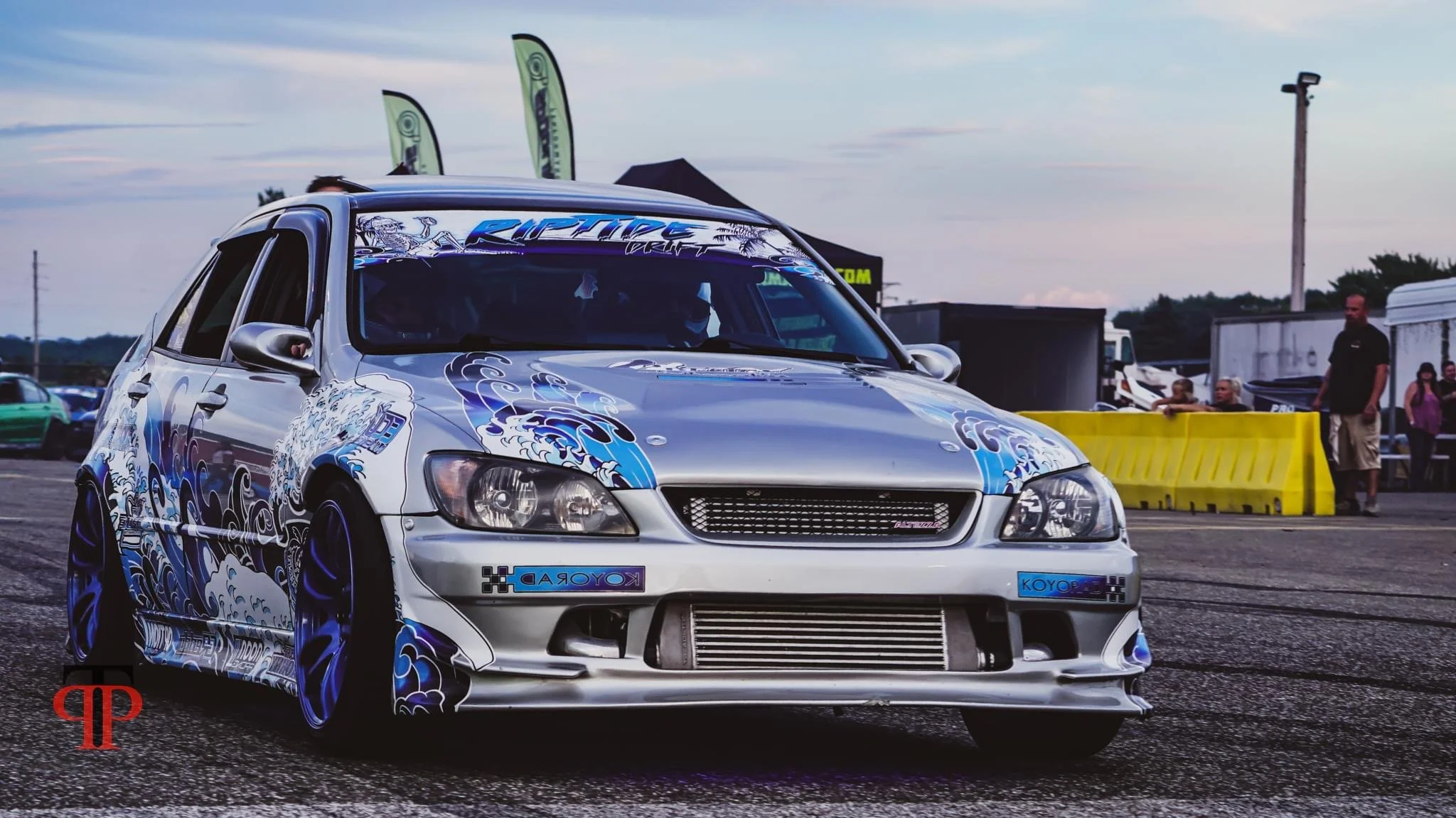A modified silver hatchback car with blue and black graphics, racing decals, and a front-mounted intercooler, parked at a car meet or race event on an asphalt lot during sunset.