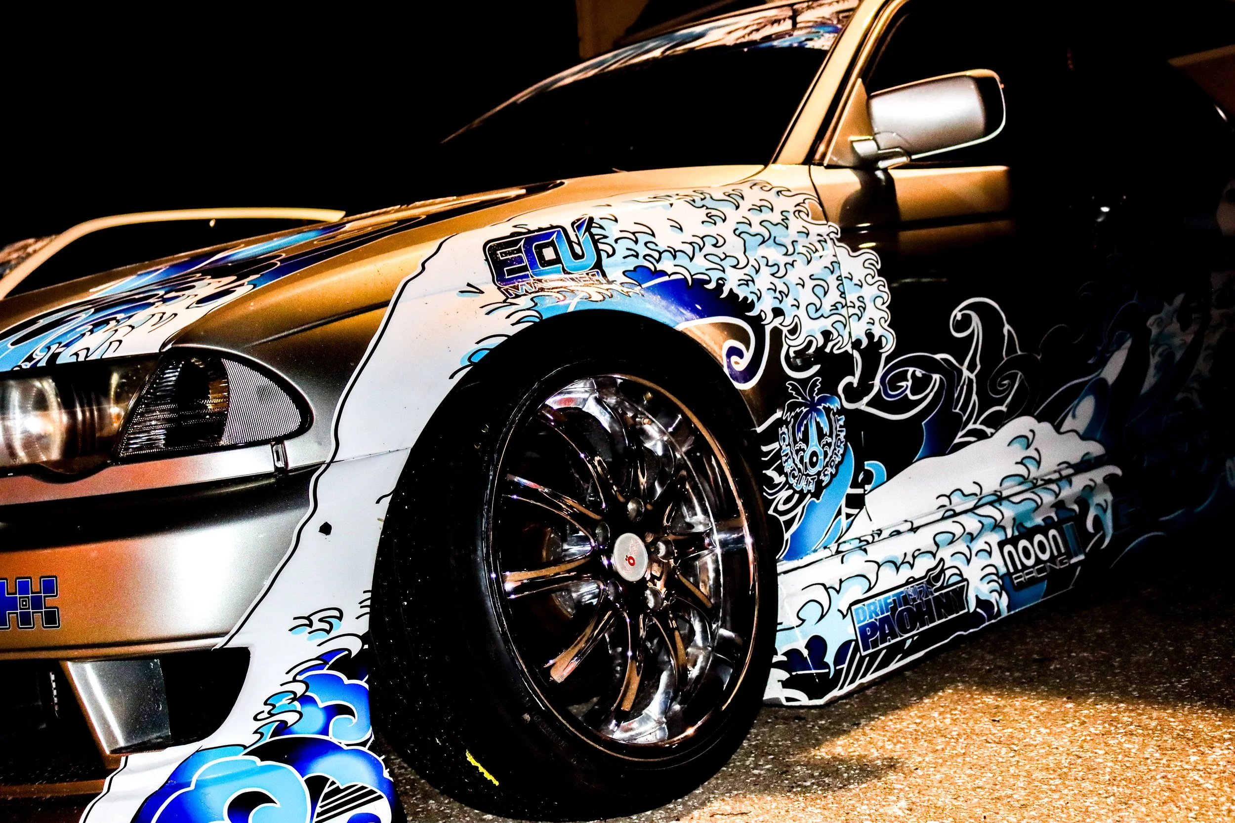 Close-up of a silver sports car with a graphic wrap featuring blue and white waves and clouds, with sponsor logos including 'ECU', 'Noon', and 'Drifting Pachinky', parked on a dark street at night.
