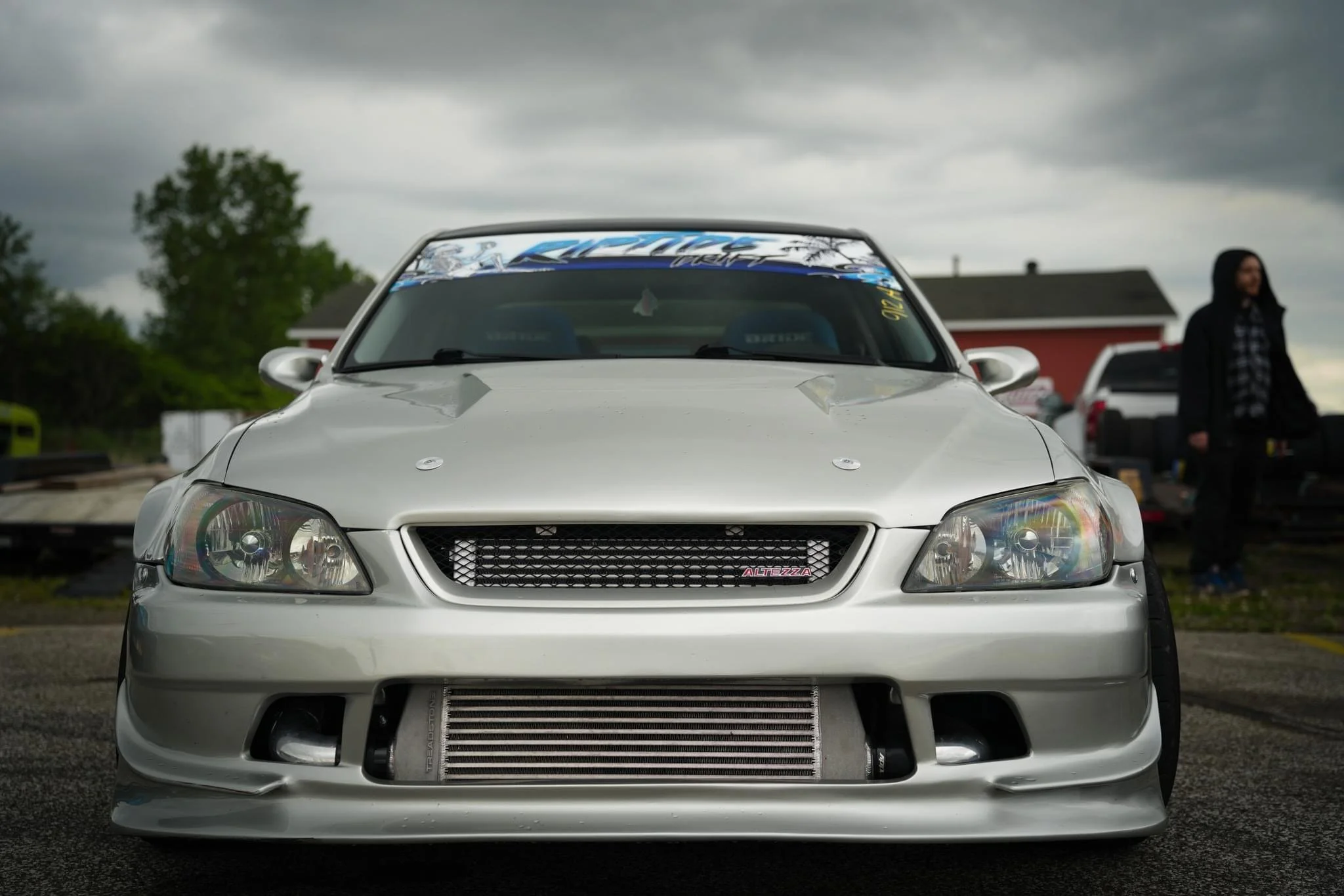 Front view of a silver modified sports car with a large intercooler, custom grille, and lowered stance, parked outdoors under a cloudy sky.
