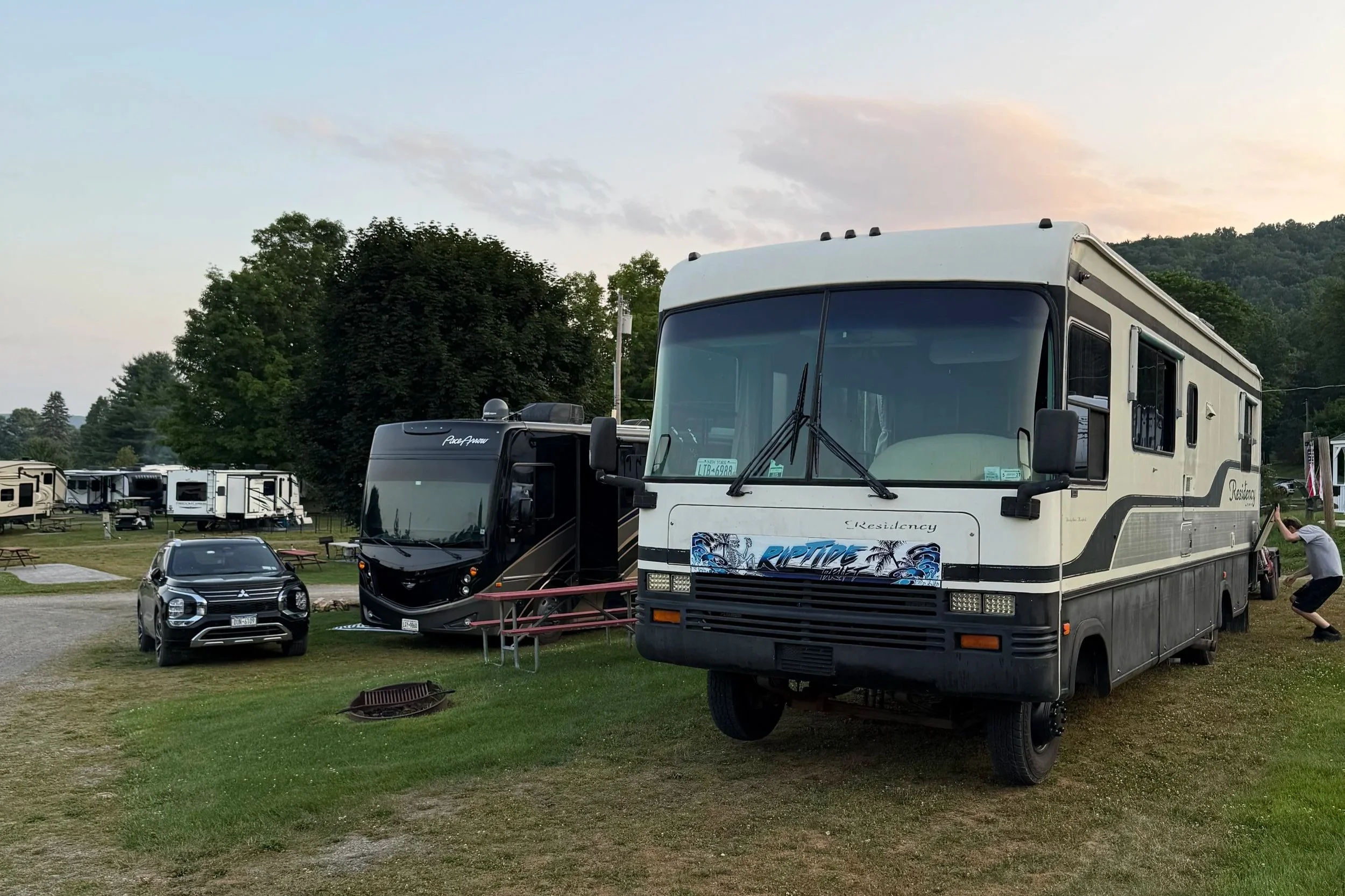 An RV park with three RVs parked on grass, a black SUV, and a person crouching near the RV on the right. There are trees, other RVs, and a picnic table in the background at dusk.