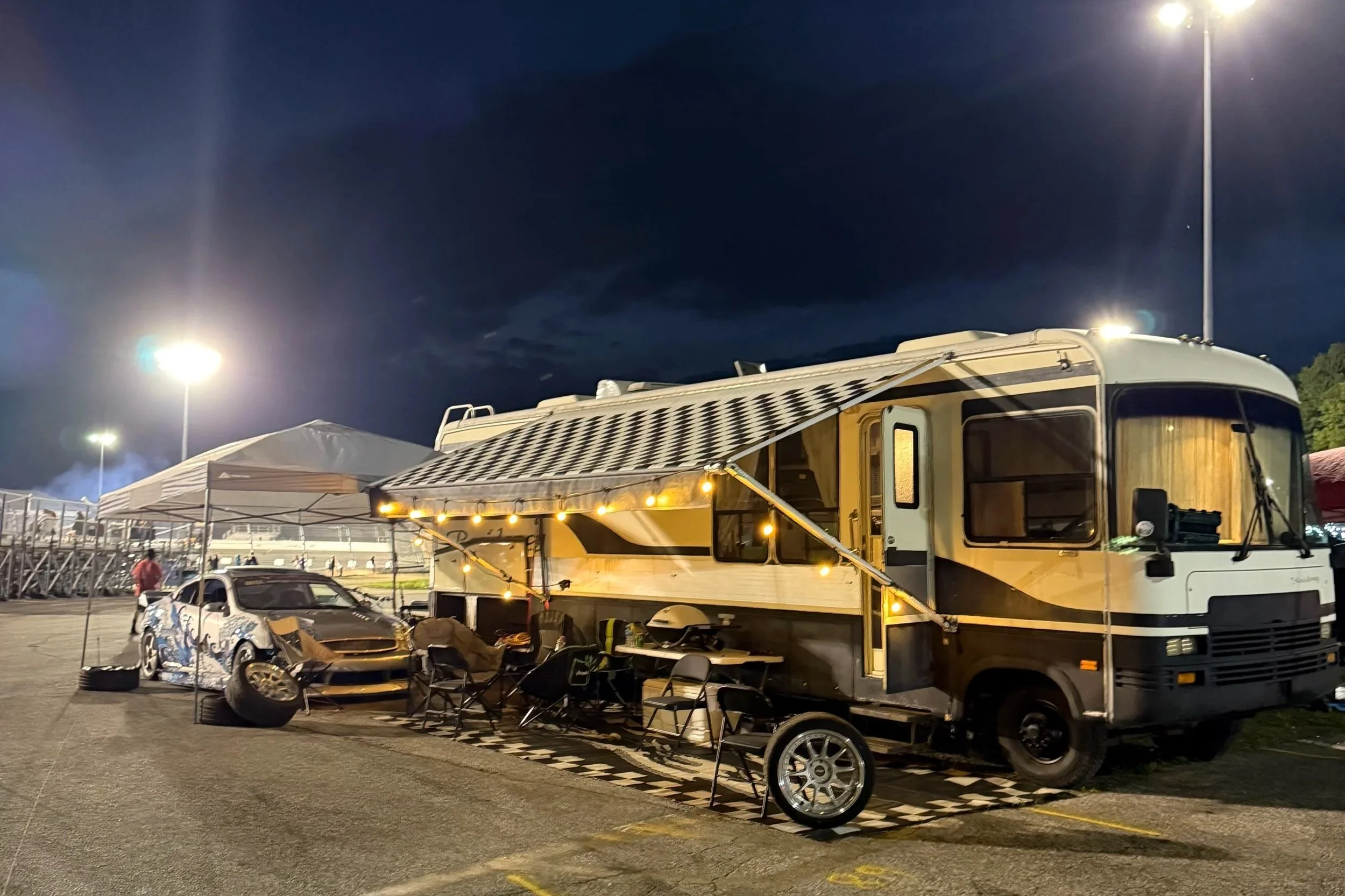 A recreational vehicle parked in a lot at night with a large awning decorated with string lights. A silver sports car with damage and a spare tire is parked nearby. There's a white canopy tent and a person in a red jacket walking in the background. The sky is dark and the area is lit by tall streetlights.