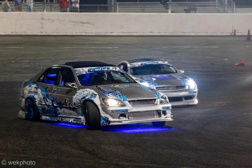 Two drift racing cars with blue underglow lights on a race track at night, one in front of the other, with a wall and spectators in the background.