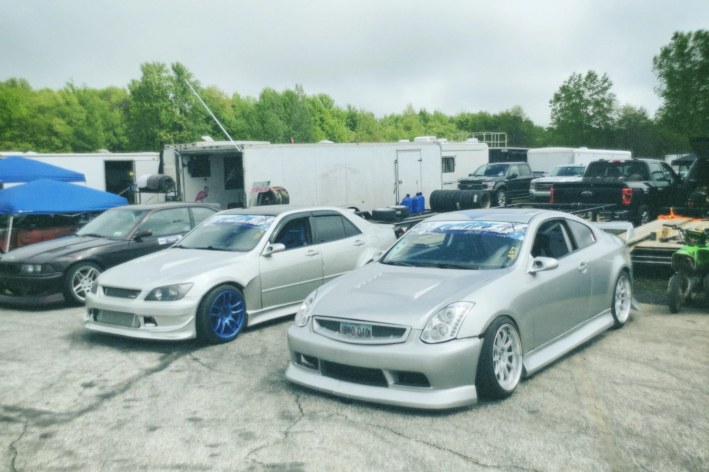Two silver sports cars with lowered suspensions and custom wheels parked side by side in an outdoor racing lot, with other vehicles and trailers in the background.