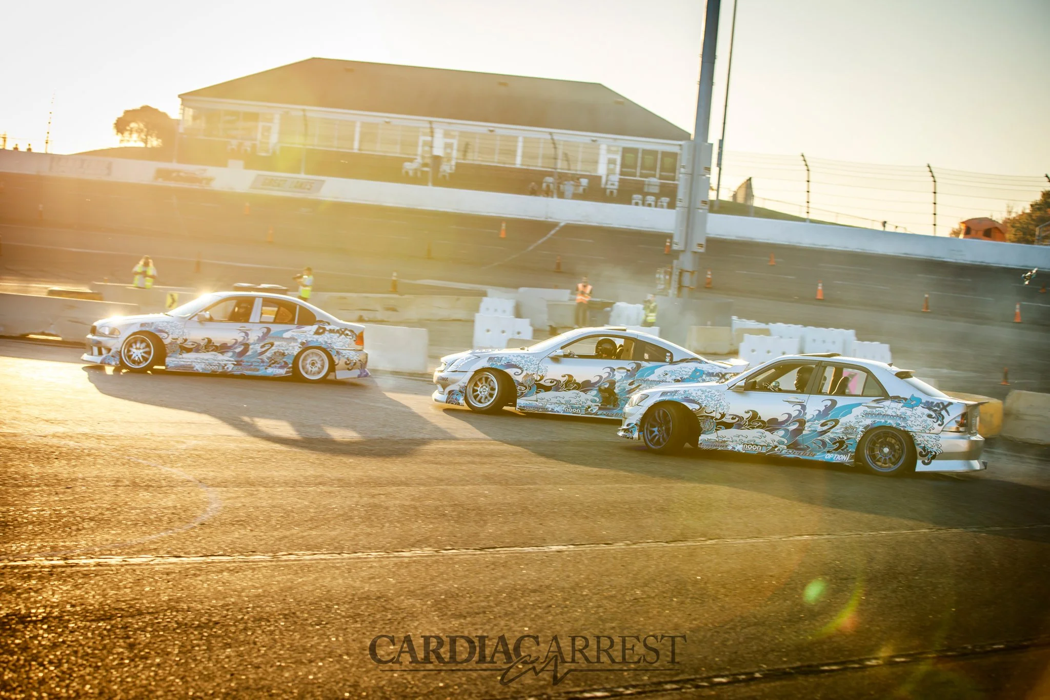 Three race cars with decorative graphics on a racetrack during sunset, with safety barriers and officials in the background.