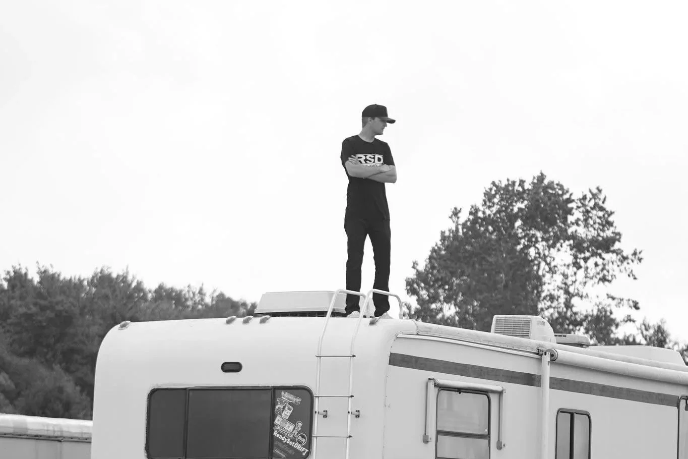 A man stands on top of a recreational vehicle with his arms crossed, looking to the side. The RV has a small window and an air conditioning unit on top. The background features trees and a clear sky.