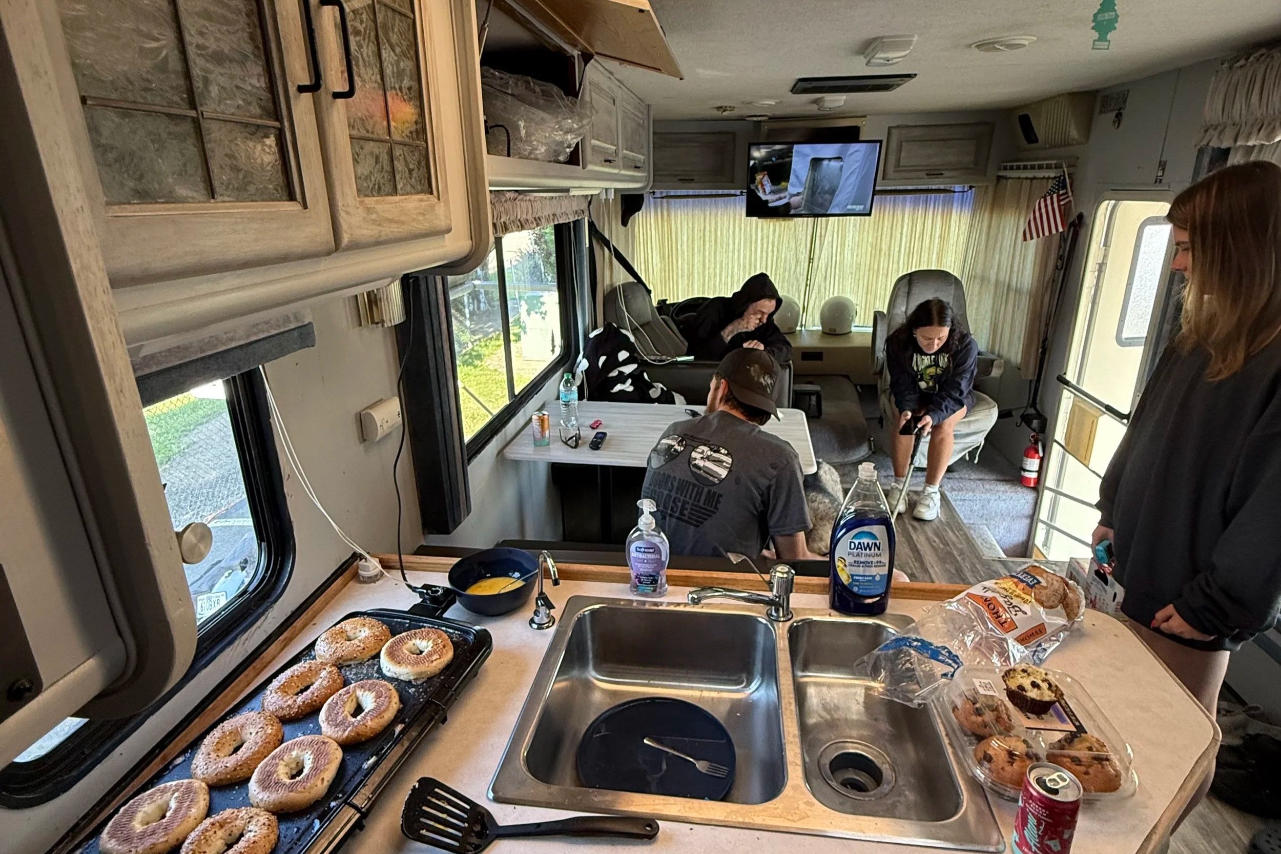Inside a camper with a kitchen counter filled with bagels, baked goods, and drinks. People are sitting and standing in the living area, some using phones, with a television on the wall, windows, and a door leading outside.