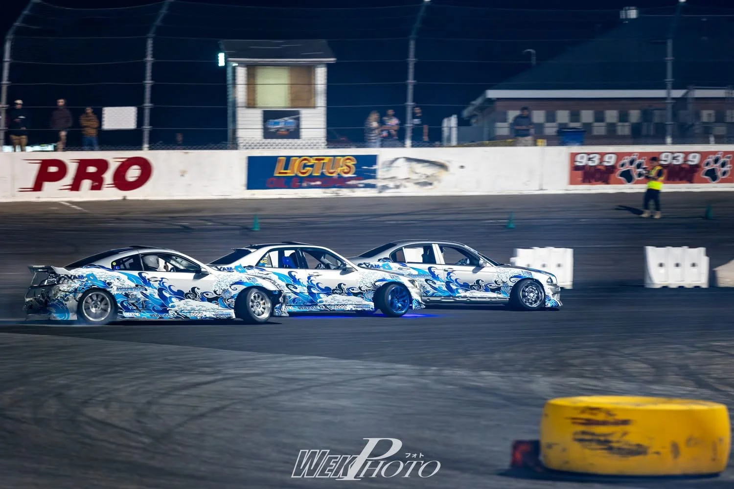 Two racing cars with blue and white wave designs on a track at night, with spectators and officials in the background.